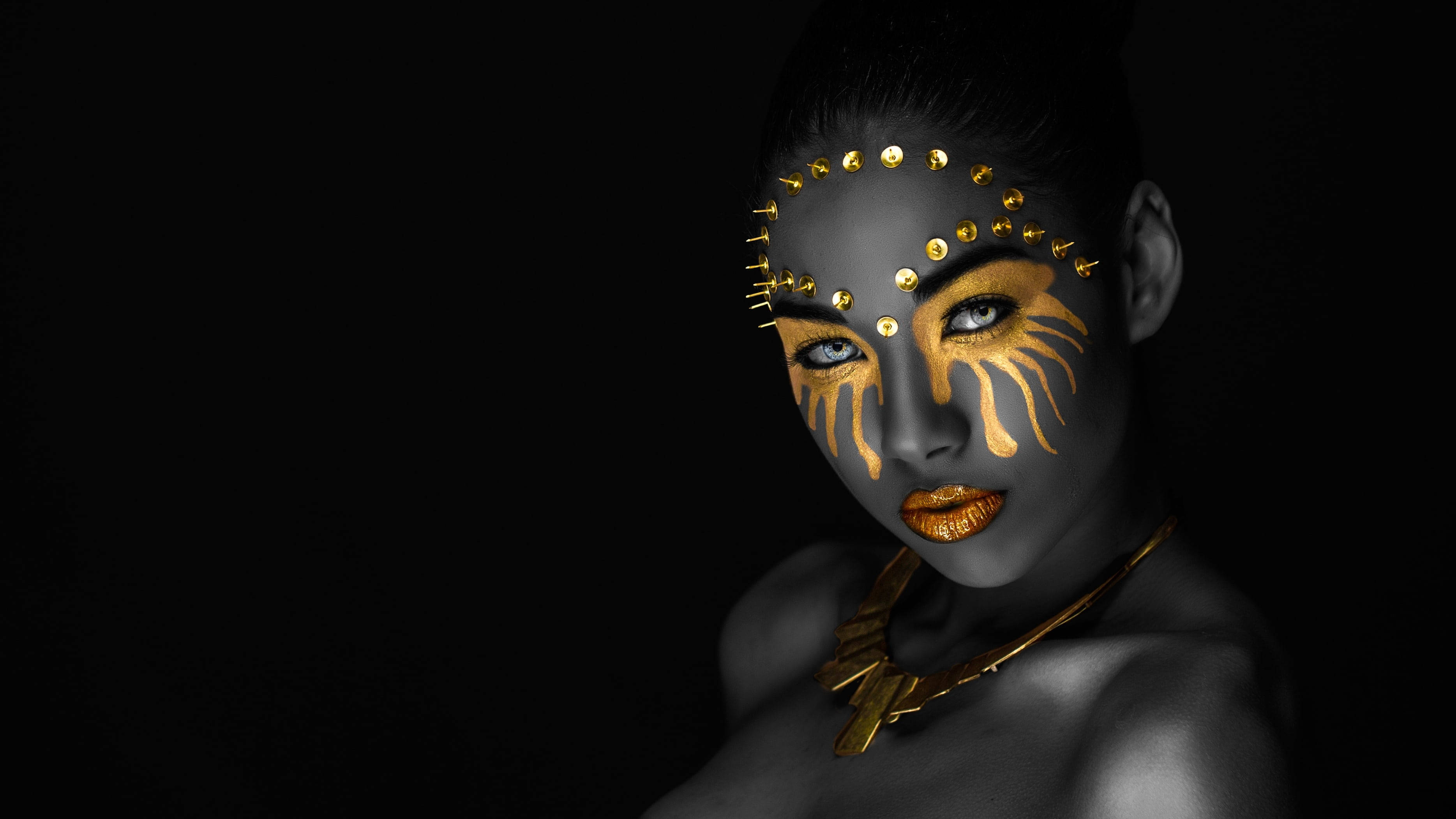 Beautiful Woman 桌布 壁紙 — 一位深色皮膚、臉上塗有金色顏料並飾有金屬釘的女性，在黑色背景下凝視著。