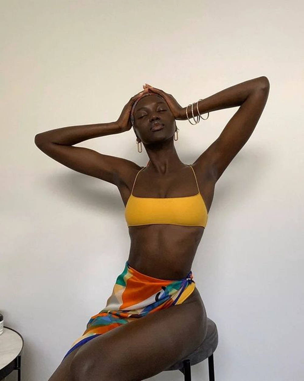 bikini model 桌布 壁紙 — 一位深色皮膚的女性坐在凳子上，身穿黃色上衣和彩色裹身裙，雙手放在腦後，背景是純白牆壁。