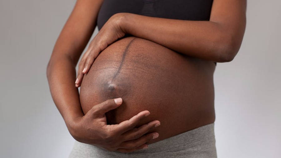 Pregnant 桌布 壁紙 — 一位深色皮膚的孕婦在柔和的灰色背景前輕輕地抱著她圓潤的肚子。