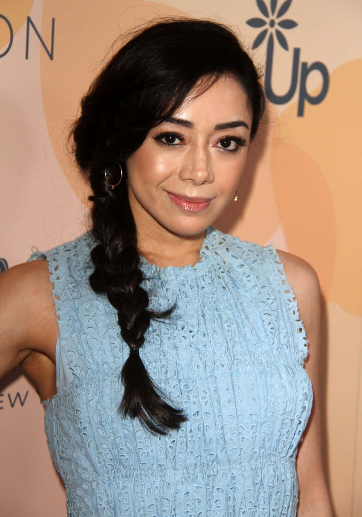Aimee Garcia 桌布 壁紙 — 一位深色辮子女性在溫暖橙色背景的活動中，穿著淺藍色蕾絲連衣裙微笑。