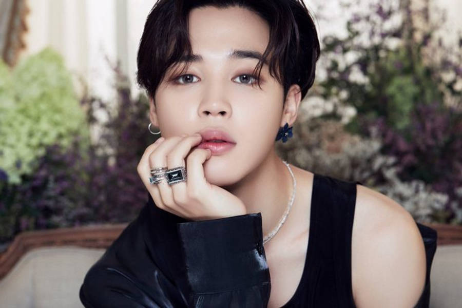 Bts Jimin 桌布 壁紙 — 一位深色頭髮、佩戴大膽珠寶的年輕男子，在茂盛的花卉背景前若有所思地擺姿勢。