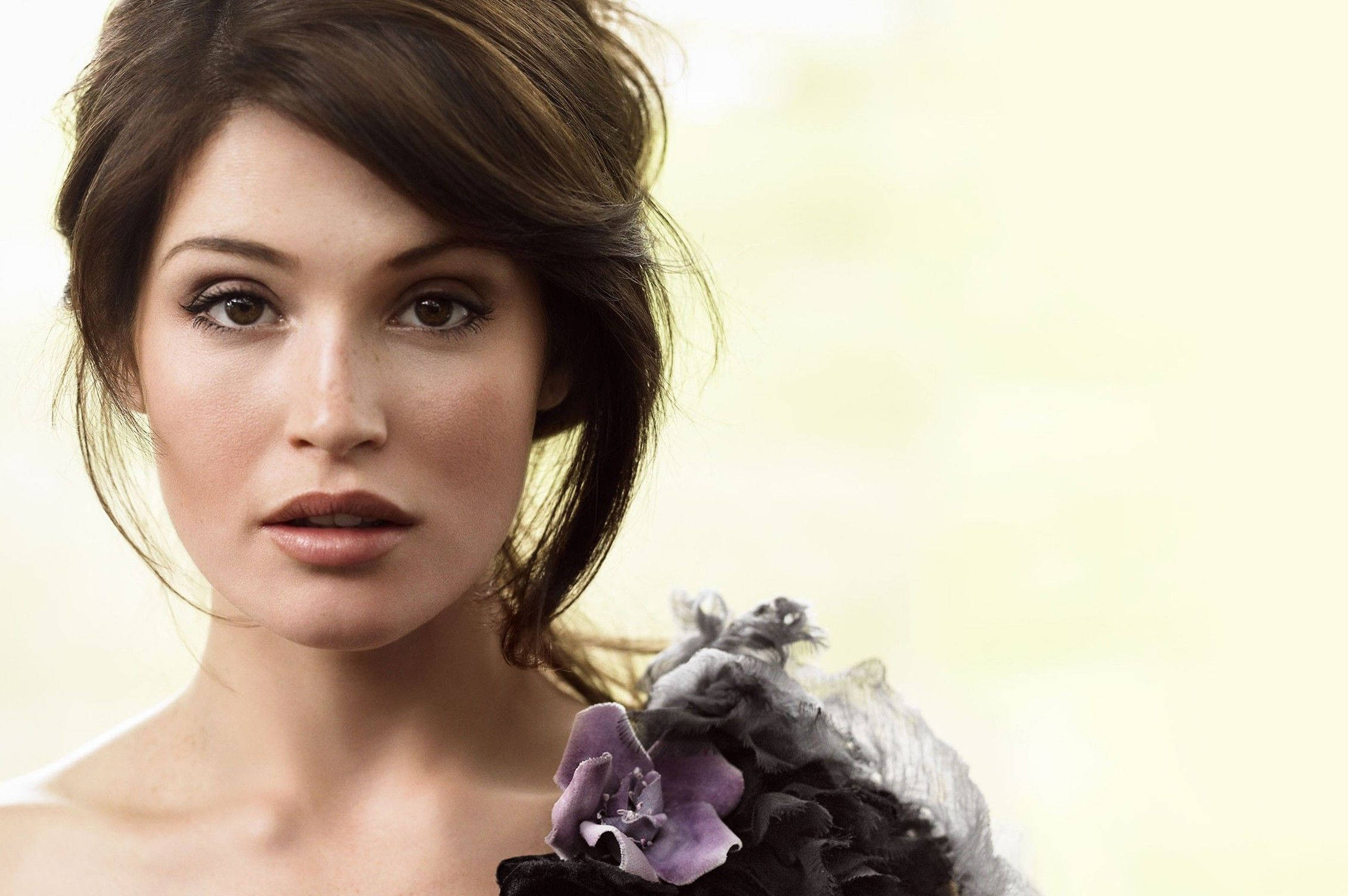 Gemma Arterton 桌布 壁紙 — 一位深色頭髮、佩戴紫色花卉配飾的女性，在柔和的黃色背景下的特寫肖像。