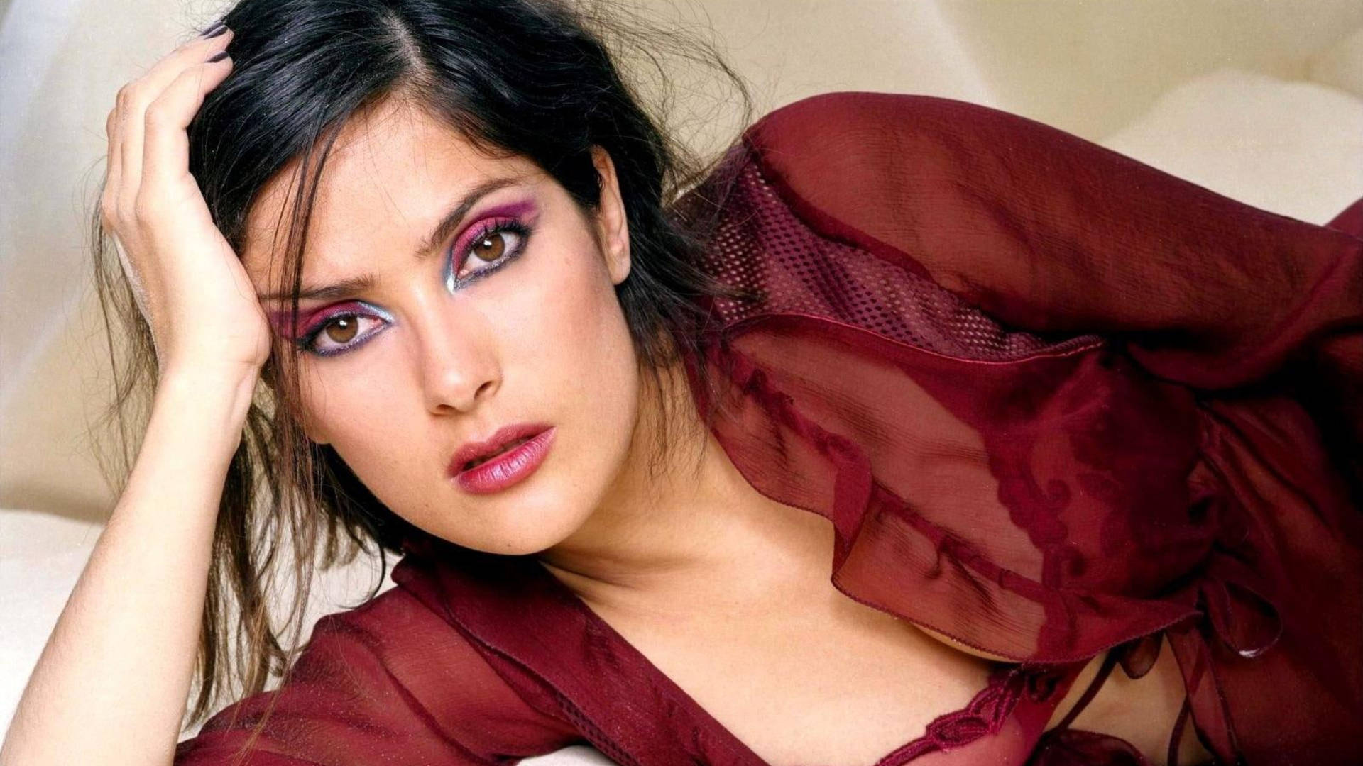 Salma 桌布 壁紙 — 一位深色頭髮、化著大膽紫色眼妝的女性，將頭靠在手上，披著一件薄透的栗色衣服，凝視著前方。