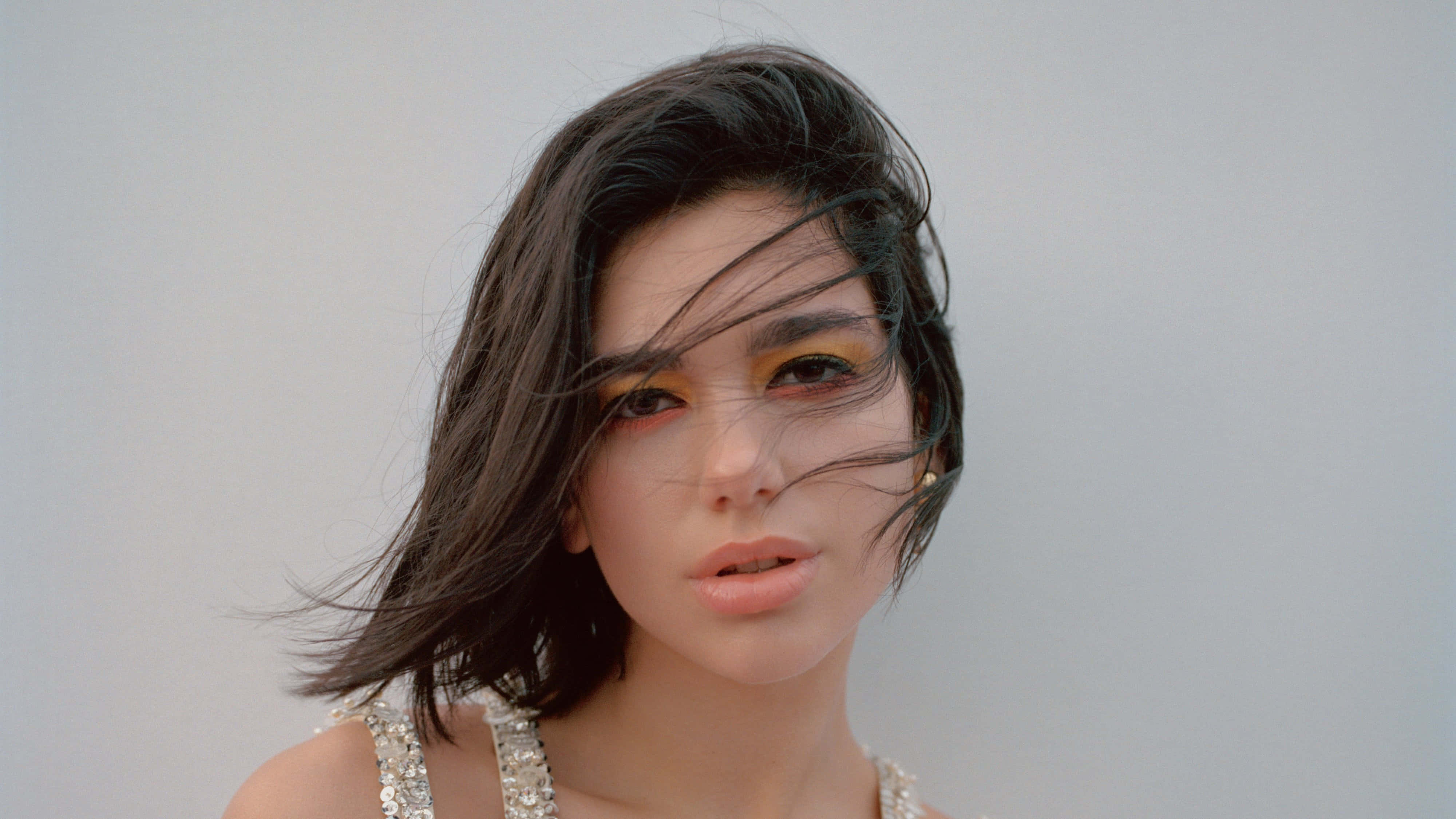 Dua Lipa 桌布 壁紙 — 一位深色頭髮、化著大膽黃色眼妝的女性，在純灰色背景下，風吹動髮絲掠過臉龐，她凝視著鏡頭。