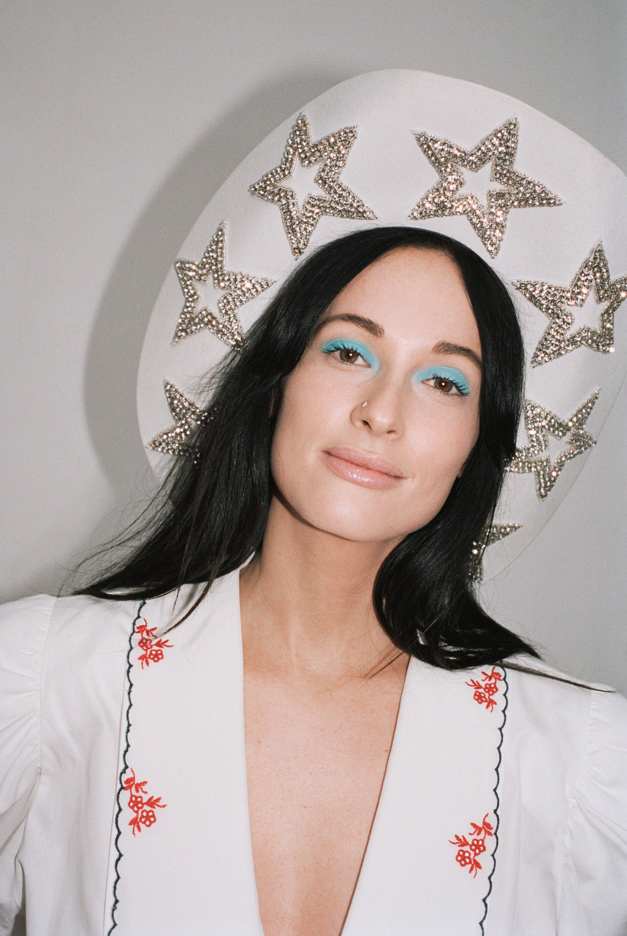 Kacey Musgraves 桌布 壁紙 — 一位深色頭髮、塗上亮藍色眼影的女性，戴著一頂飾有閃爍星星的白色帽子，身穿帶有紅色刺繡的白色襯衫。