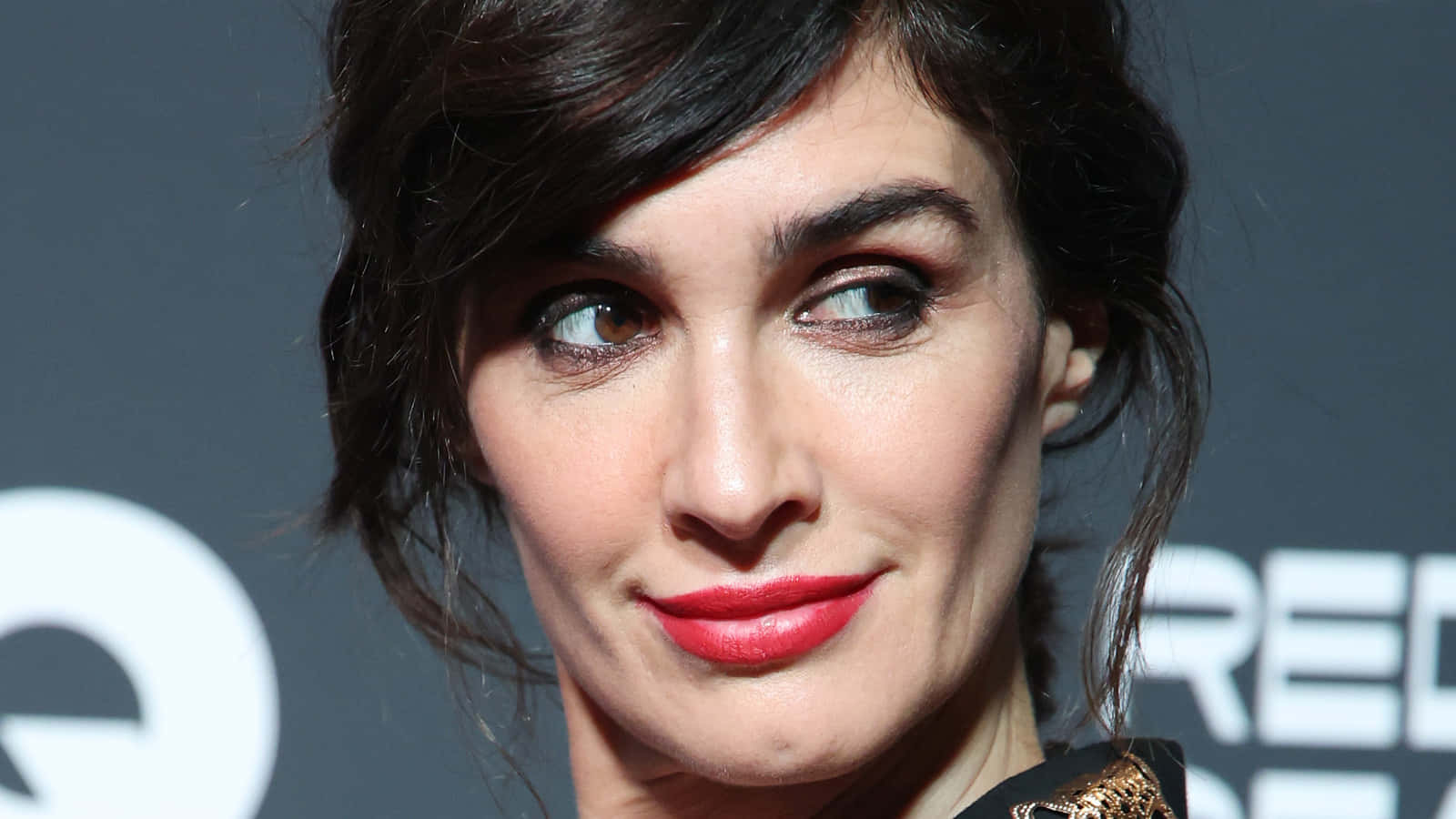 Paz Vega 桌布 壁紙 — 一位深色頭髮、塗著大膽紅色唇膏的女性，帶著微妙微笑望向別處的特寫肖像。