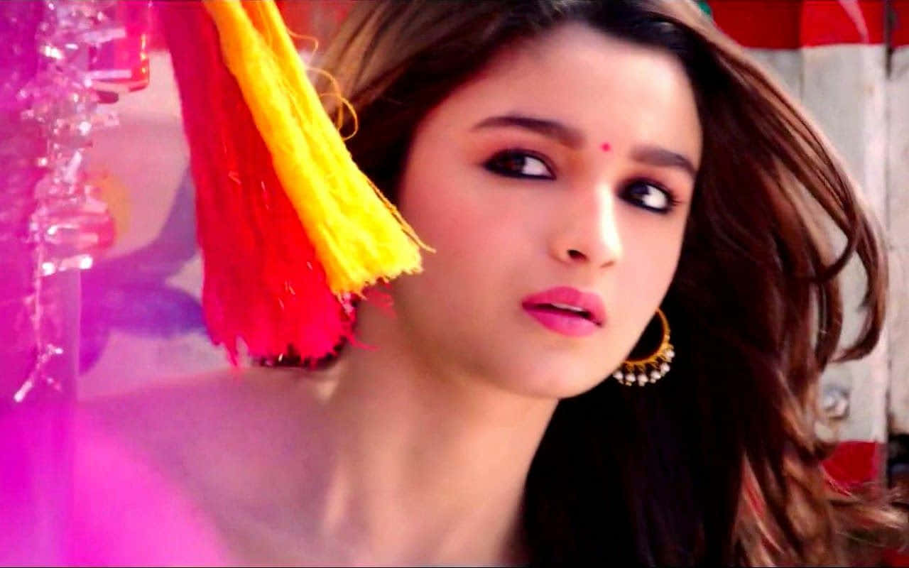 Alia Bhatt 桌布 壁紙 — 一位深色頭髮、塗著粉紅色口紅的女性，在粉紅色燈光下，在鮮豔的紅色和黃色流蘇旁邊凝視著。