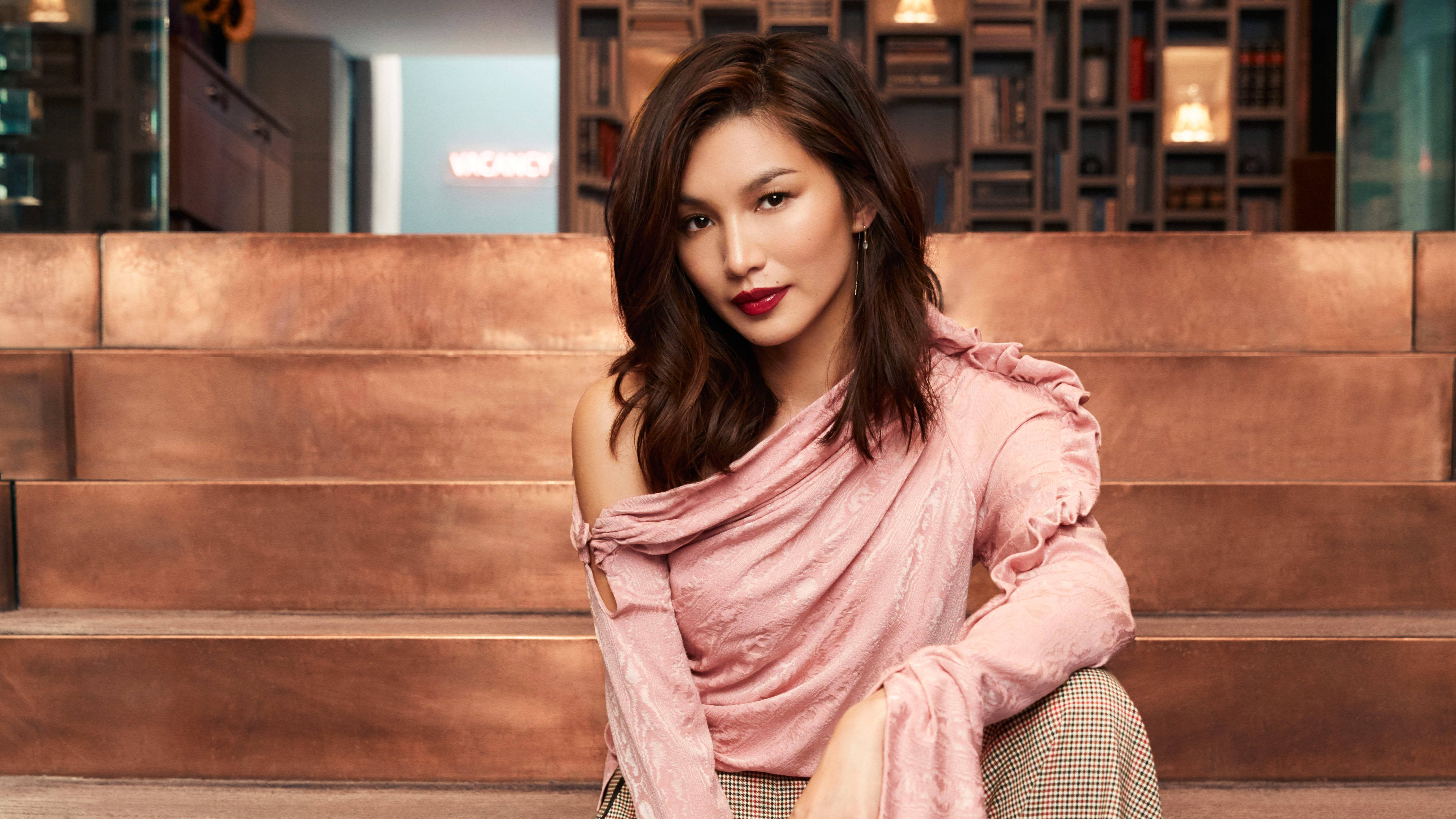 Gemma Chan 桌布 壁紙 — 一位深色頭髮、塗著紅色唇膏的女性自信地坐在現代室內銅色調的台階上，身後有書架。