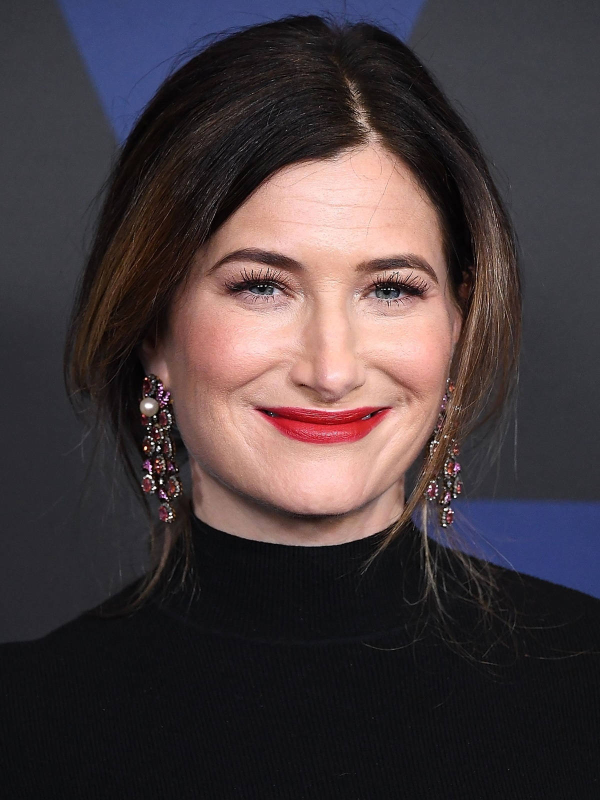 Celebrity 桌布 壁紙 — 一位深色頭髮、塗著紅色唇膏的女性，在幾何藍色和灰色背景前溫暖微笑的特寫肖像。