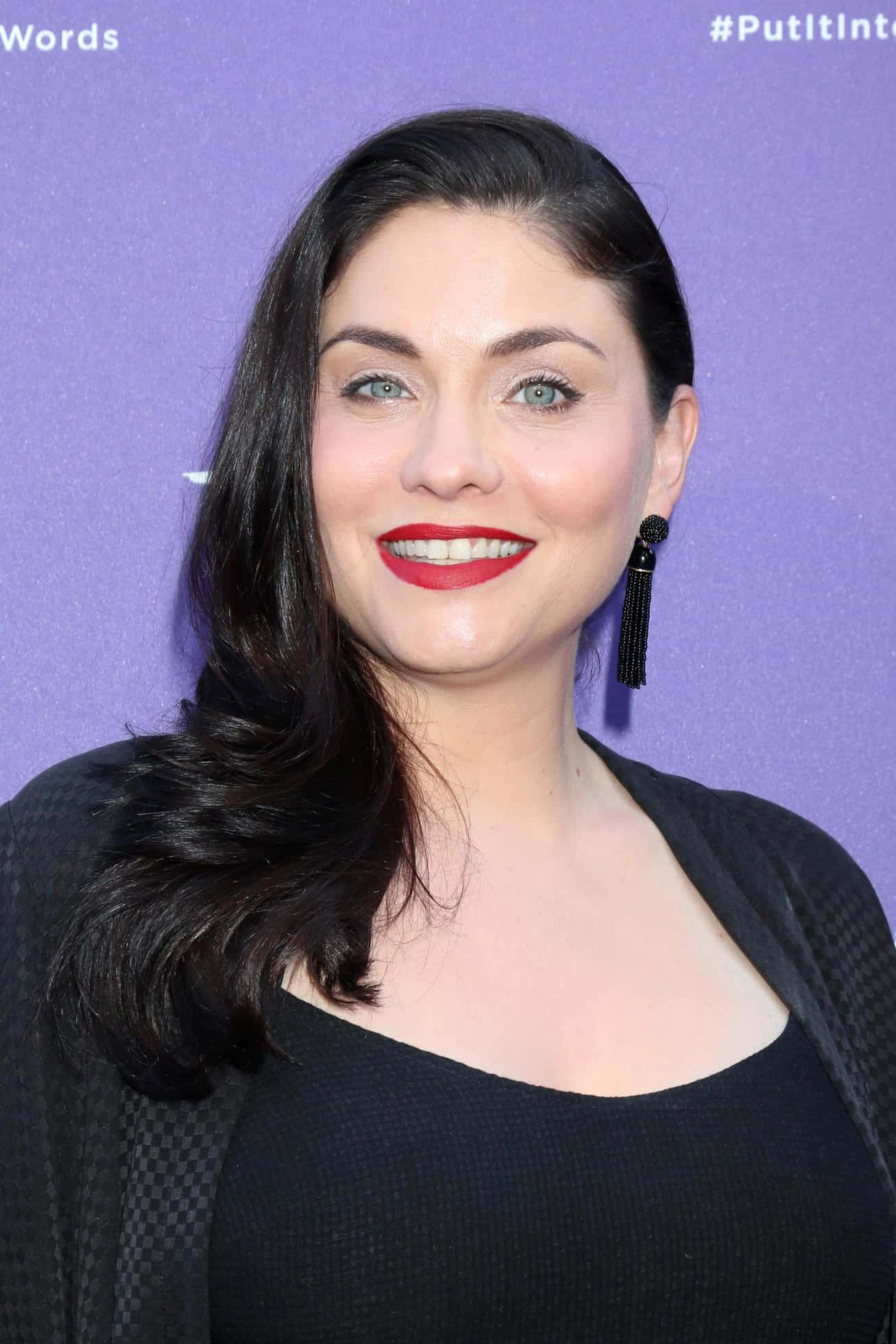 jodi lyn o'keefe 桌布 壁紙 — 一位深色頭髮、塗著紅色唇膏的女性在紫色背景的活動中微笑。