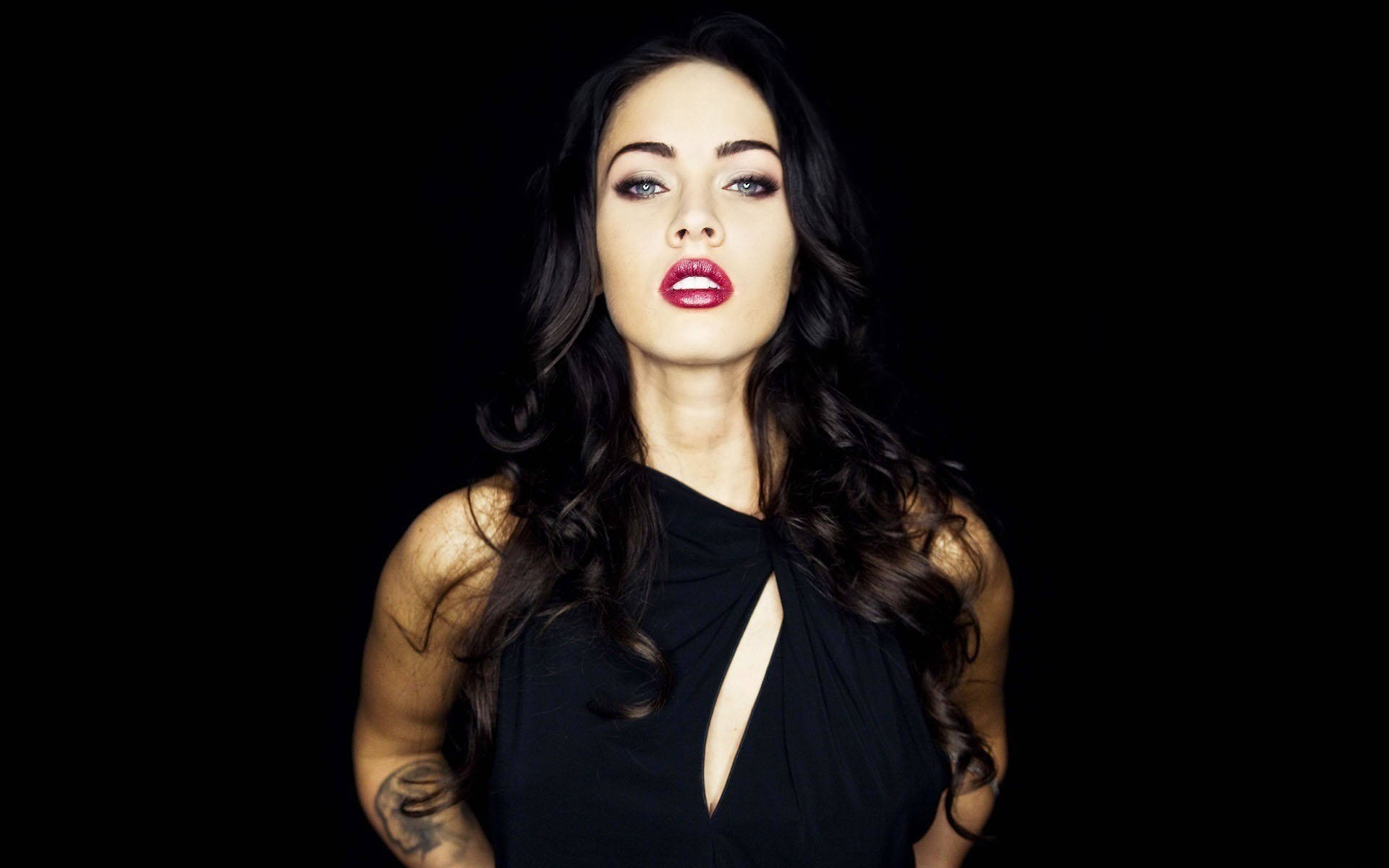 Megan Fox HD 桌布 壁紙 — 一位深色頭髮、大膽紅唇的女性在純黑色背景下強烈凝視。