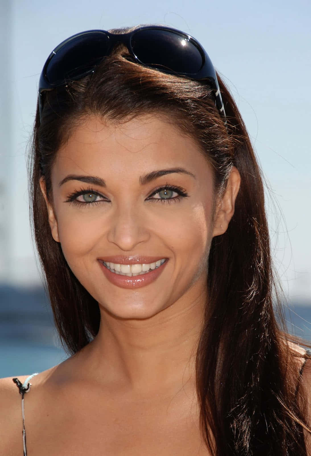Aishwarya Rai 桌布 壁紙 — 一位深色頭髮、太陽眼鏡架在頭上的女性，在明亮的藍天下溫暖地微笑。