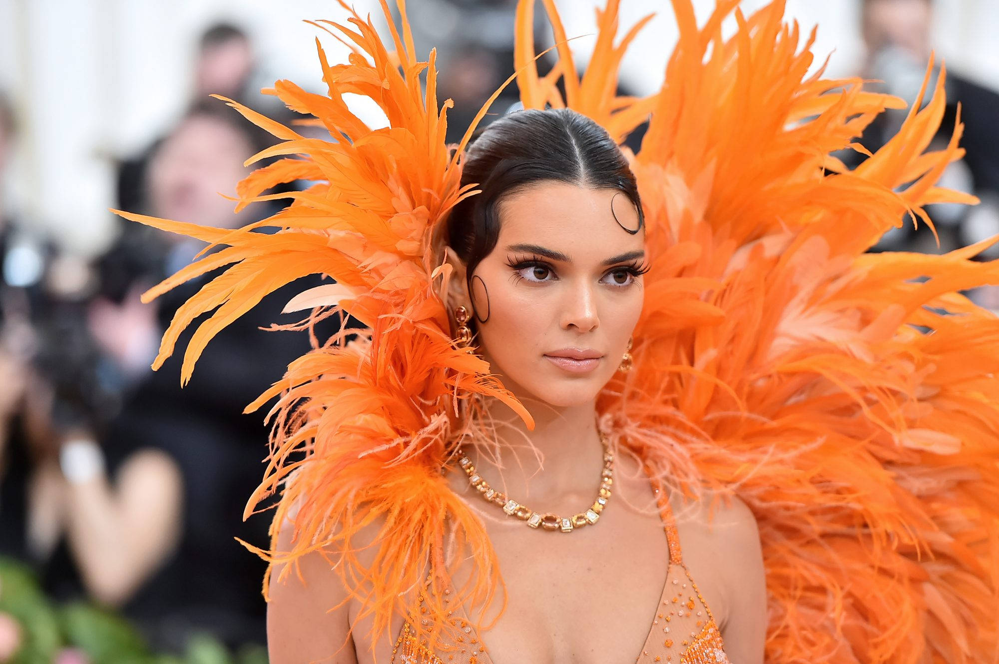 kendall 桌布 壁紙 — 一位深色頭髮、妝容大膽的女性，在一個華麗的活動中戴著戲劇性的橙色羽毛頭飾和相配的服裝。