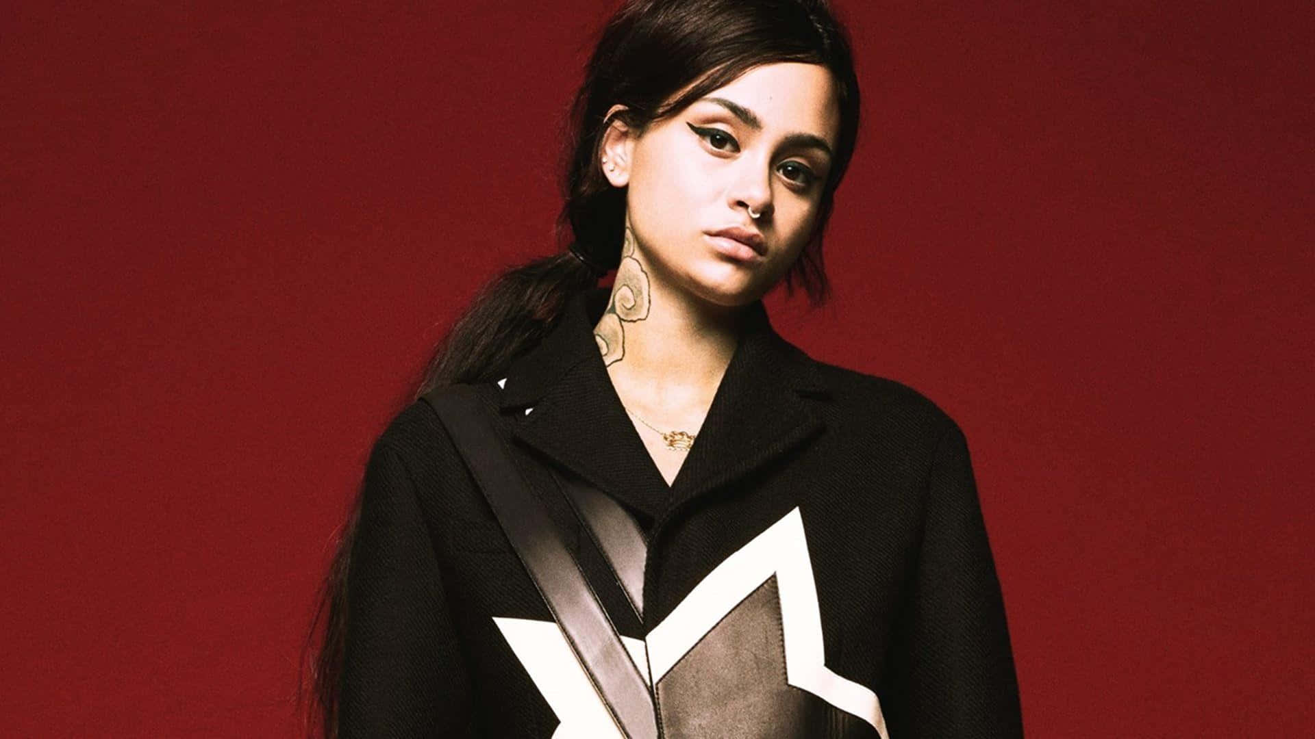 kehlani 桌布 壁紙 — 一位深色頭髮、妝容大膽的女性，在深紅色背景下穿著帶有白色幾何圖案的黑色外套。