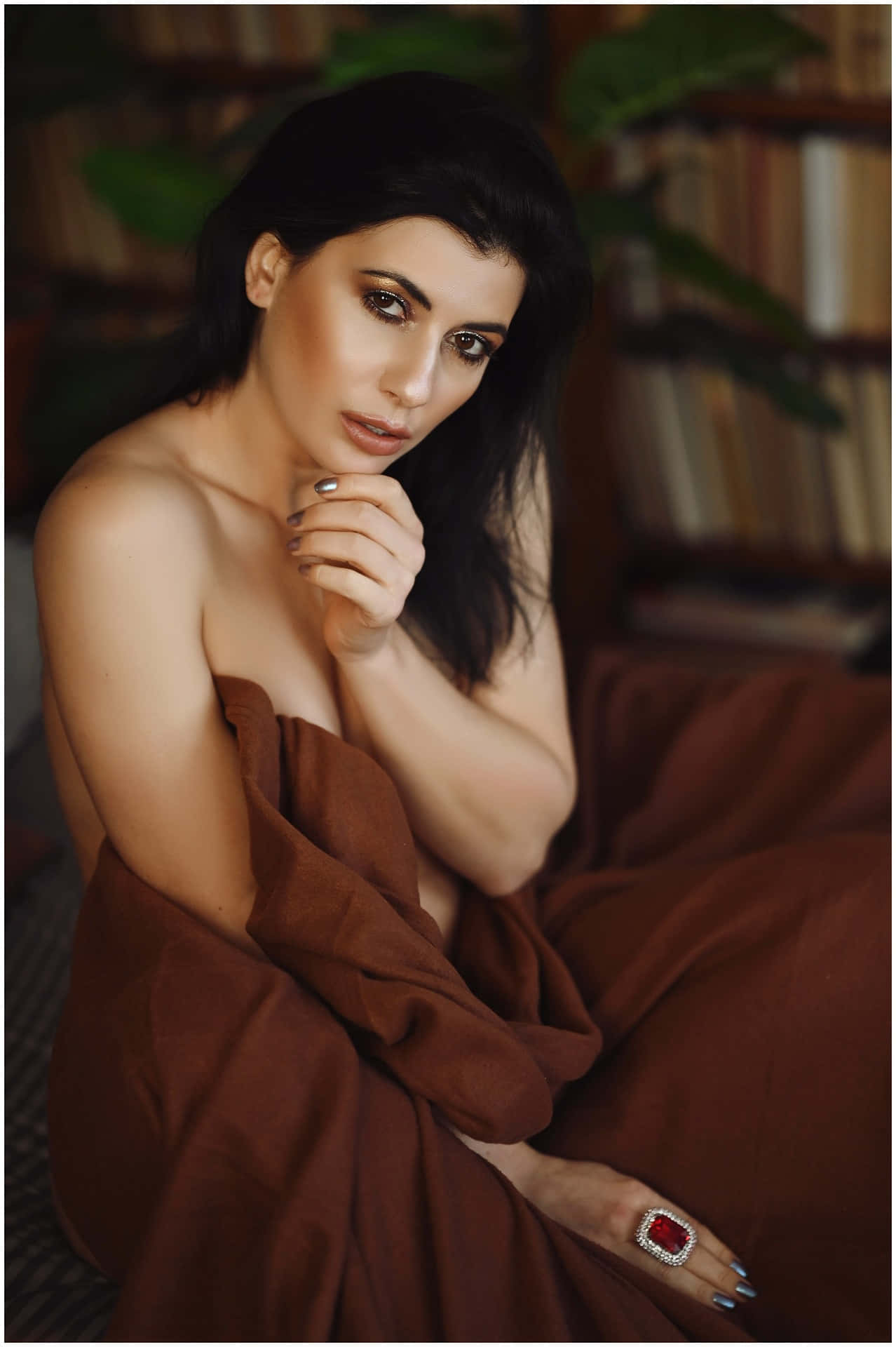 Sensual 桌布 壁紙 — 一位深色頭髮、妝容大膽的女性，披著濃郁的棕色布料坐著，戴著引人注目的紅色戒指，凝視著。