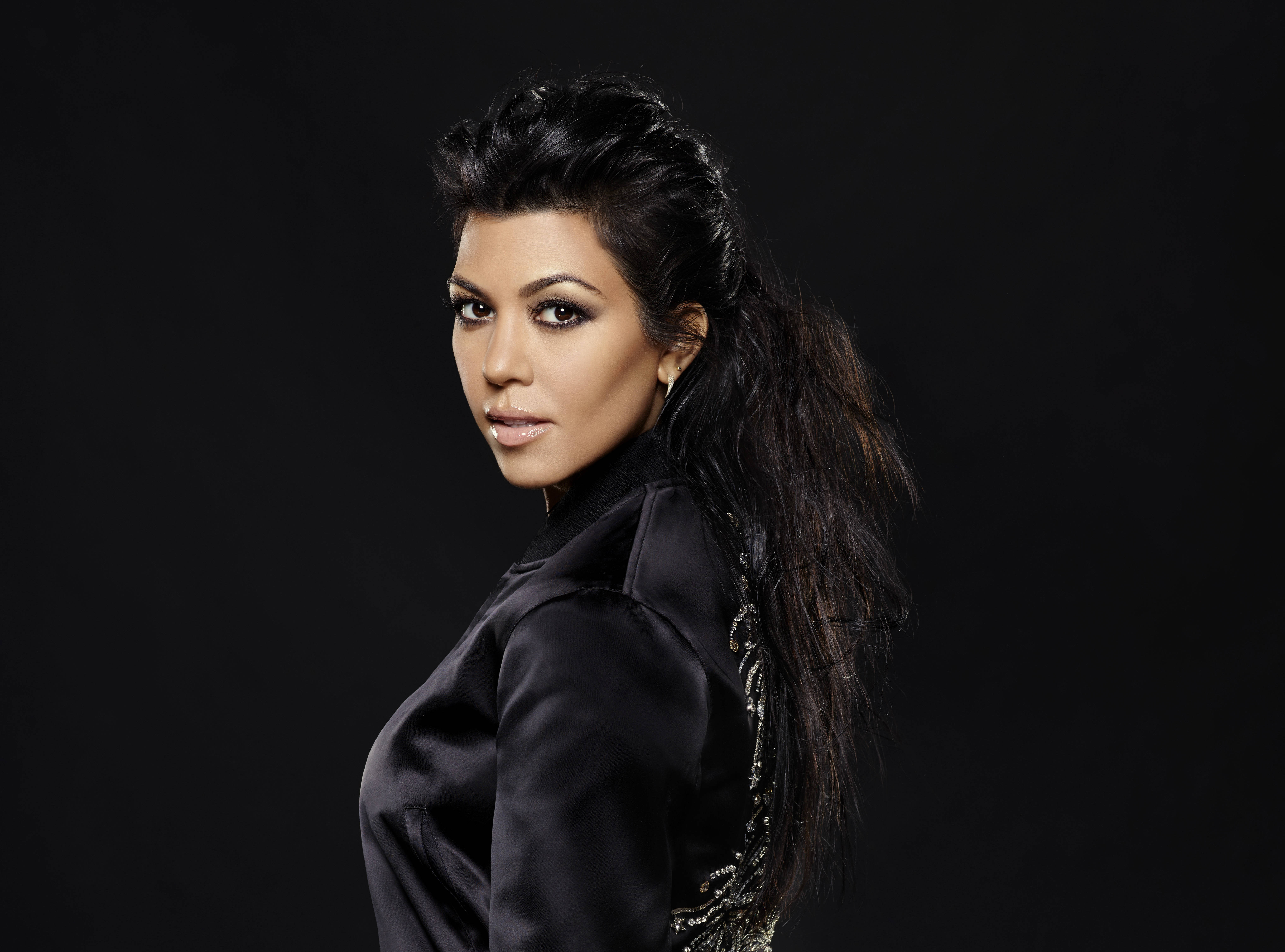 Kourtney 桌布 壁紙 — 一位深色頭髮、妝容大膽的女性，身穿黑色緞面外套，在黑色背景前回頭望向肩膀。