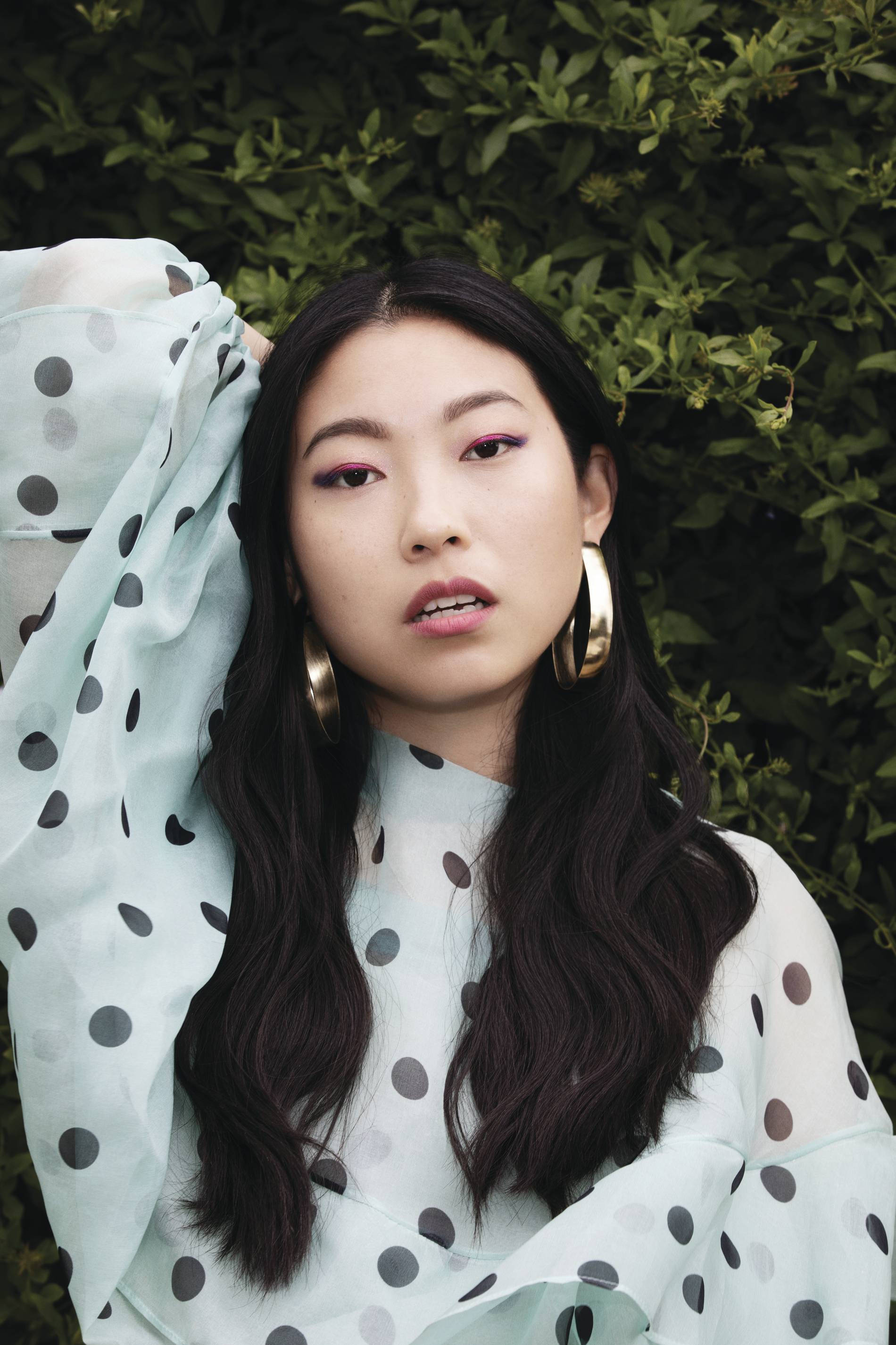 Awkwafina 桌布 壁紙 — 一位深色頭髮、妝容大膽的年輕女性，在茂密的綠色樹籬前擺姿勢，身穿淺藍色圓點襯衫和金色耳環。