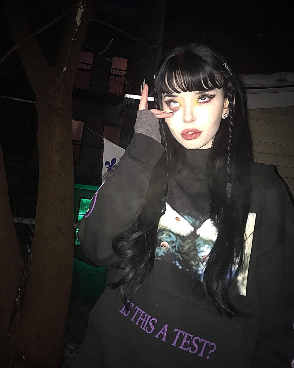 Girl Aesthetic 桌布 壁紙 — 一位深色頭髮、妝容大膽的年輕女性在夜晚昏暗的路燈下拿著香煙。