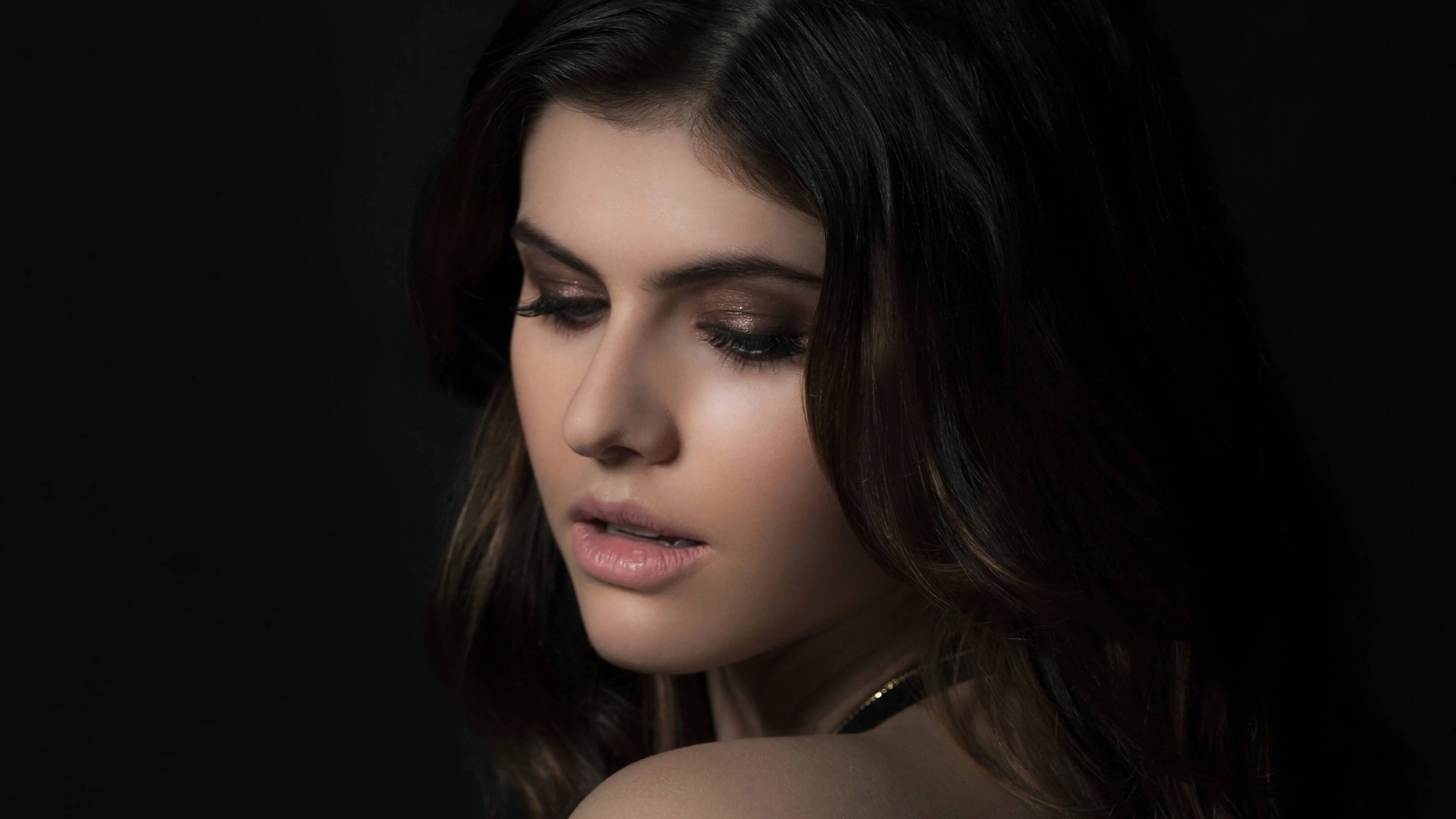 Alexandra Daddario 4K 桌布 壁紙 — 一位深色頭髮、妝容柔和的女性在黑色背景和戲劇性燈光下向下凝視。