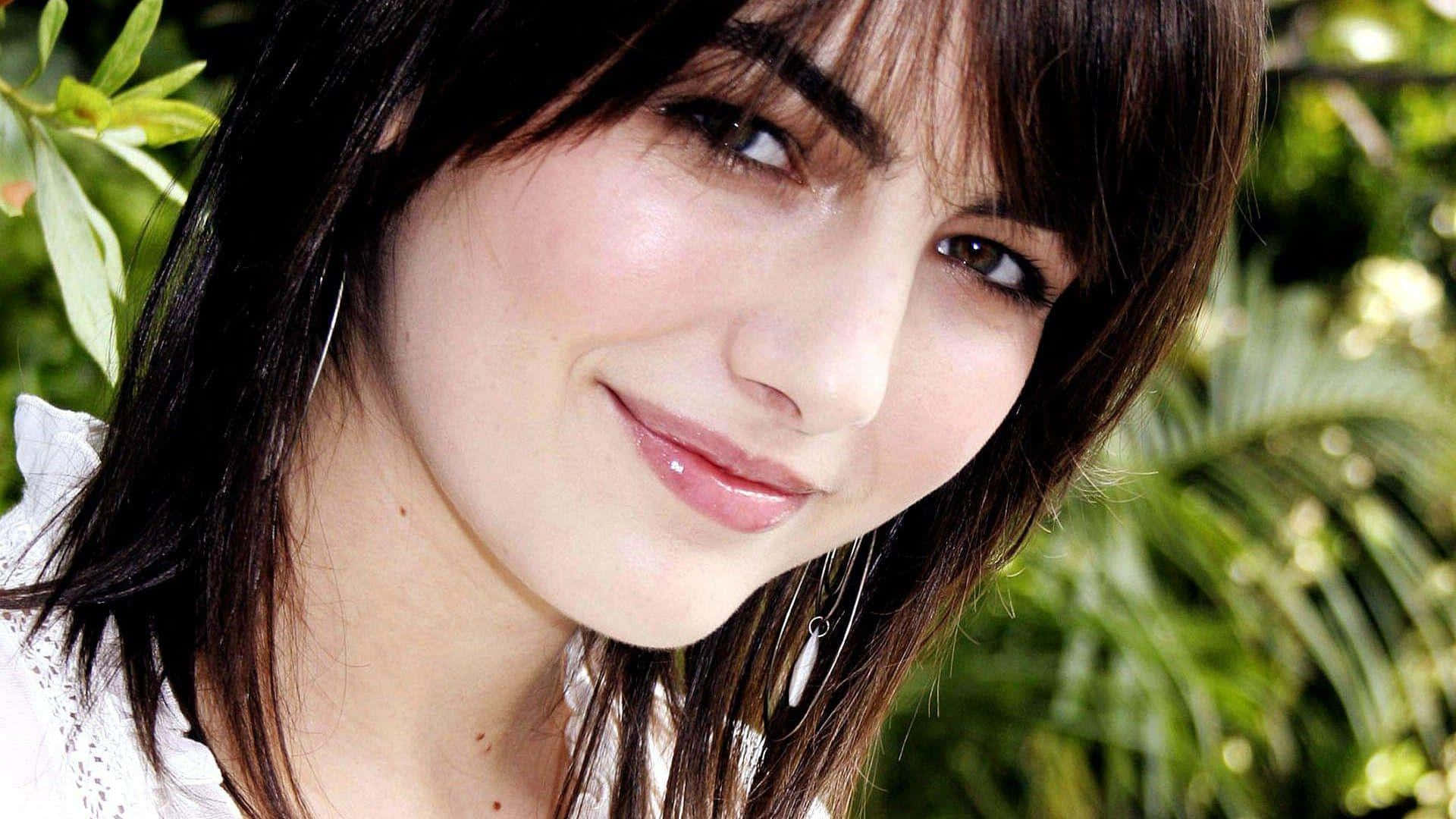 Camilla Belle 桌布 壁紙 — 一位深色頭髮、帶著溫柔微笑的女子的特寫肖像，被茂密的綠色樹葉在柔和自然光線中框住。