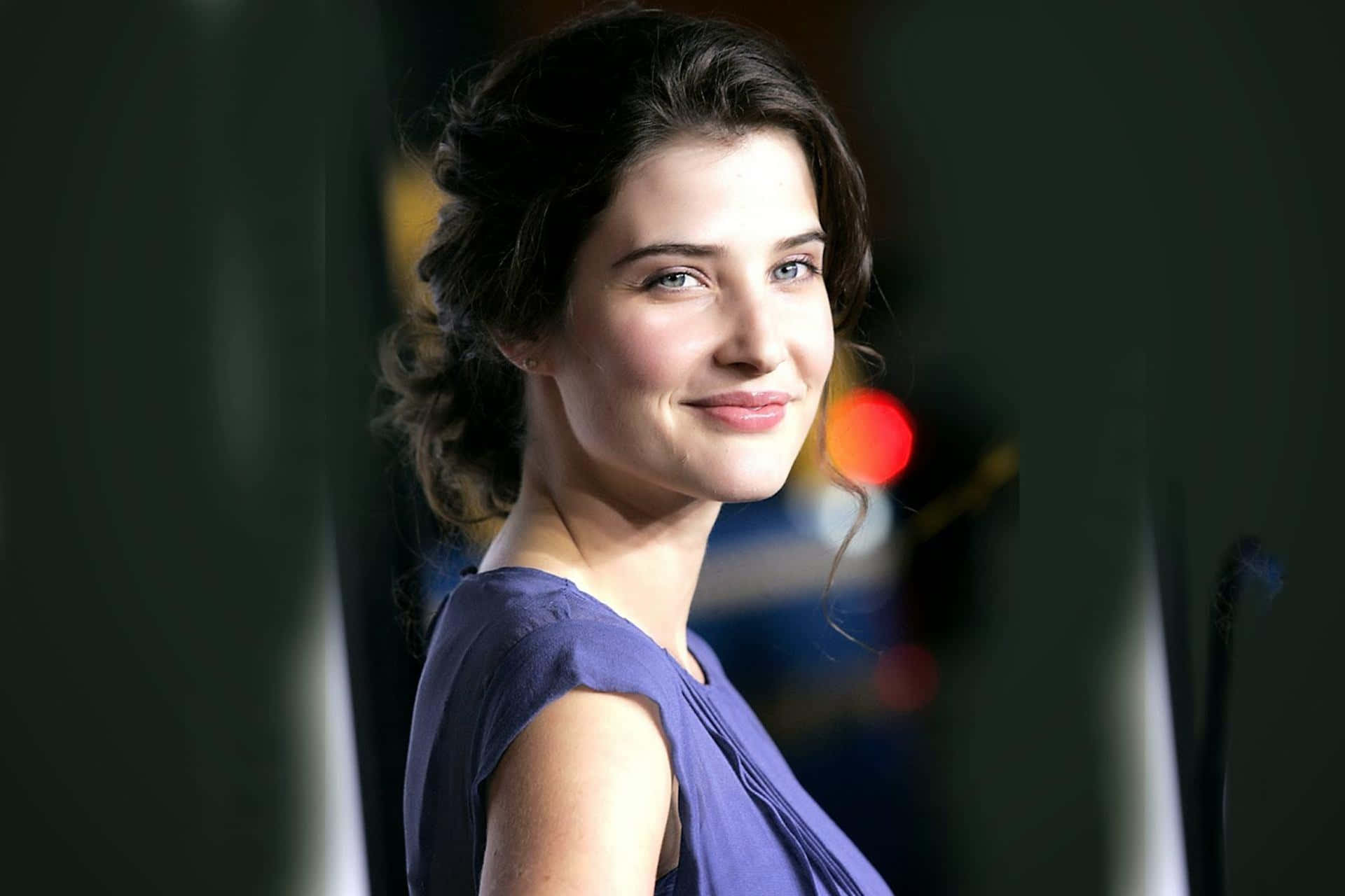 Cobie Smulders 桌布 壁紙 — 一位深色頭髮、帶著溫柔微笑的女性，穿著紫色連衣裙，背景柔和模糊，帶有溫暖的散景燈光。
