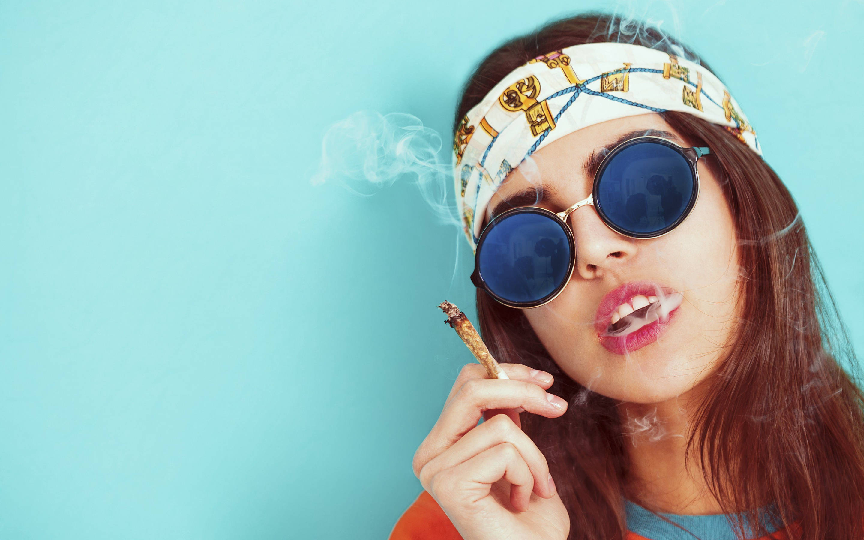 吸大麻 桌布 壁紙 — 一位深色頭髮、戴圓形太陽眼鏡的女性，在純藍色背景下手持大麻煙並呼出煙霧。