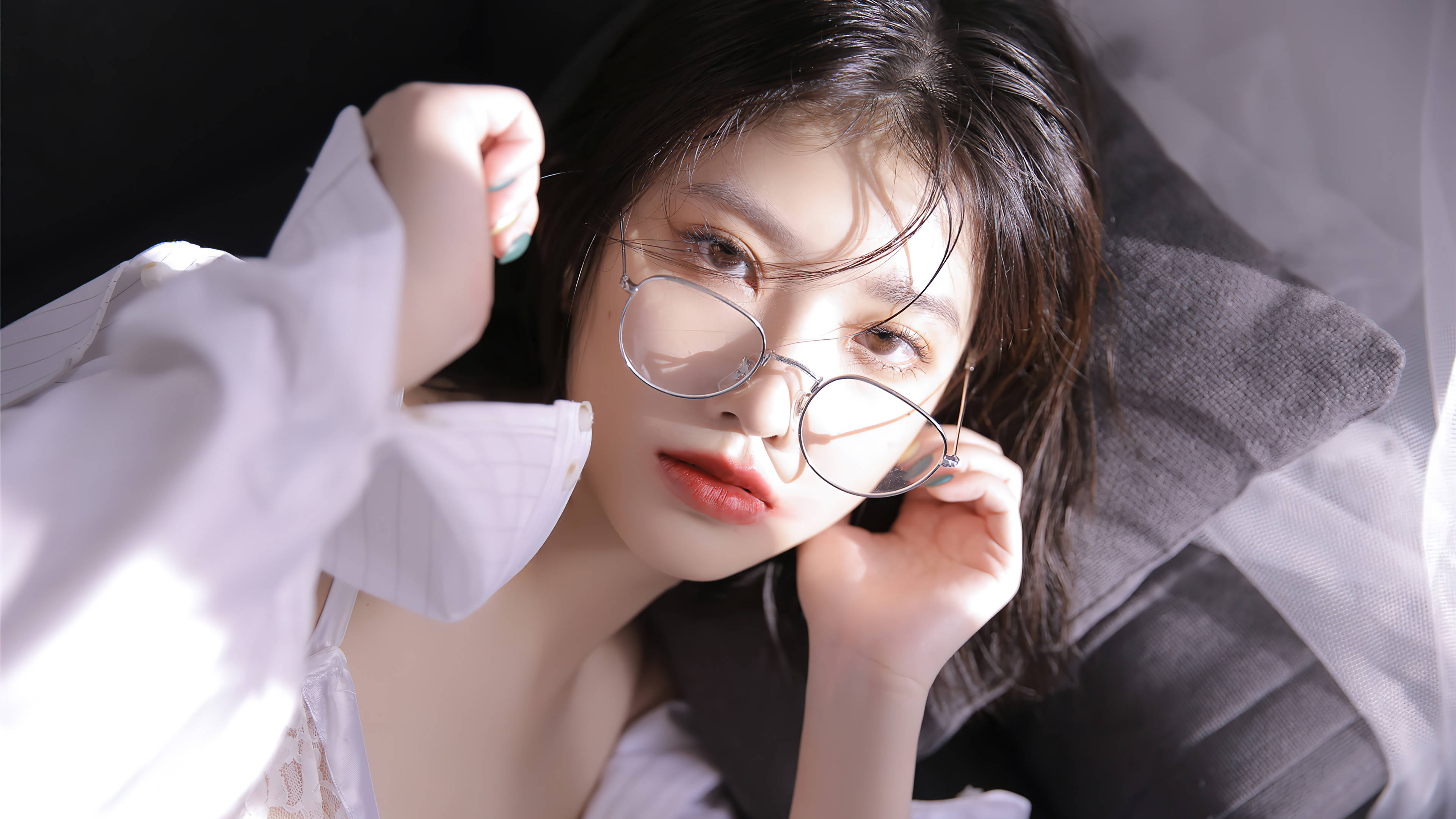 模特兒 桌布 壁紙 — 一位深色頭髮、戴圓框眼鏡的年輕女子，將頭靠在灰色枕頭上，身穿白色襯衫，在柔和光線下直視鏡頭。