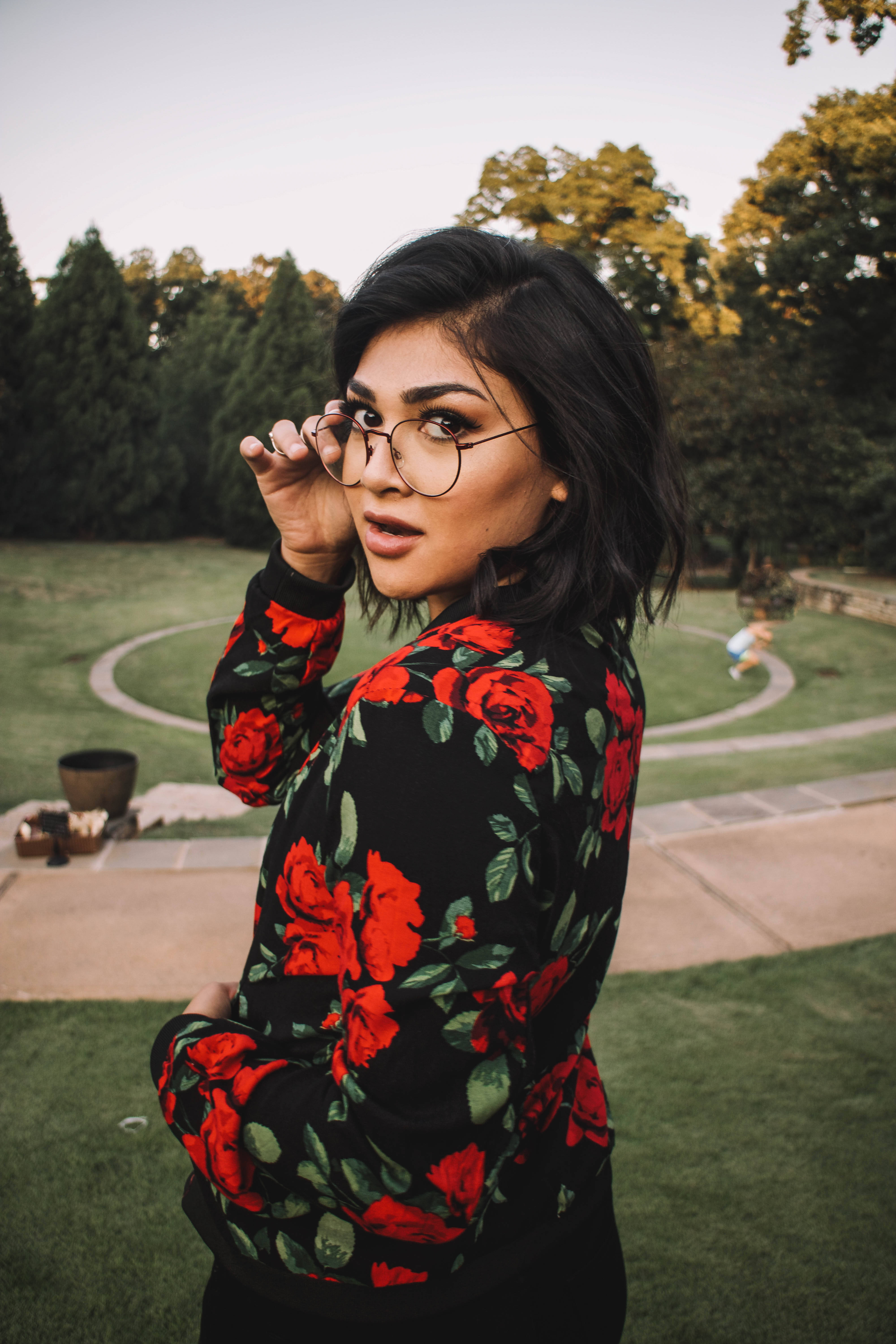 Hipster 桌布 壁紙 — 一位深色頭髮、戴眼鏡的女性，在鬱鬱蔥蔥的綠色花園中穿著帶有醒目紅色玫瑰圖案的黑色毛衣，調整她的眼鏡框。