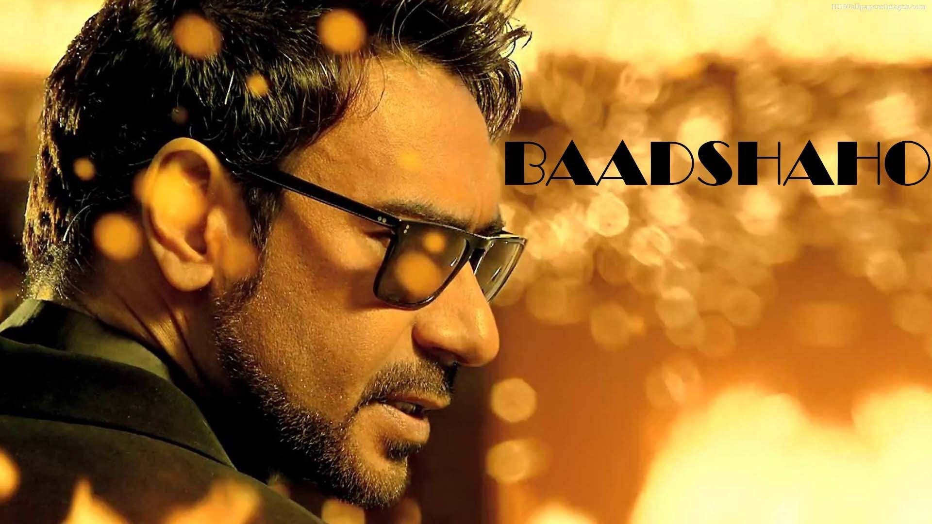 ajay devgn 桌布 壁紙 — 一位深色頭髮、戴眼鏡的男子，在溫暖的金色散景背景上疊加著「BAADSHAH」字樣，若有所思地凝視著。