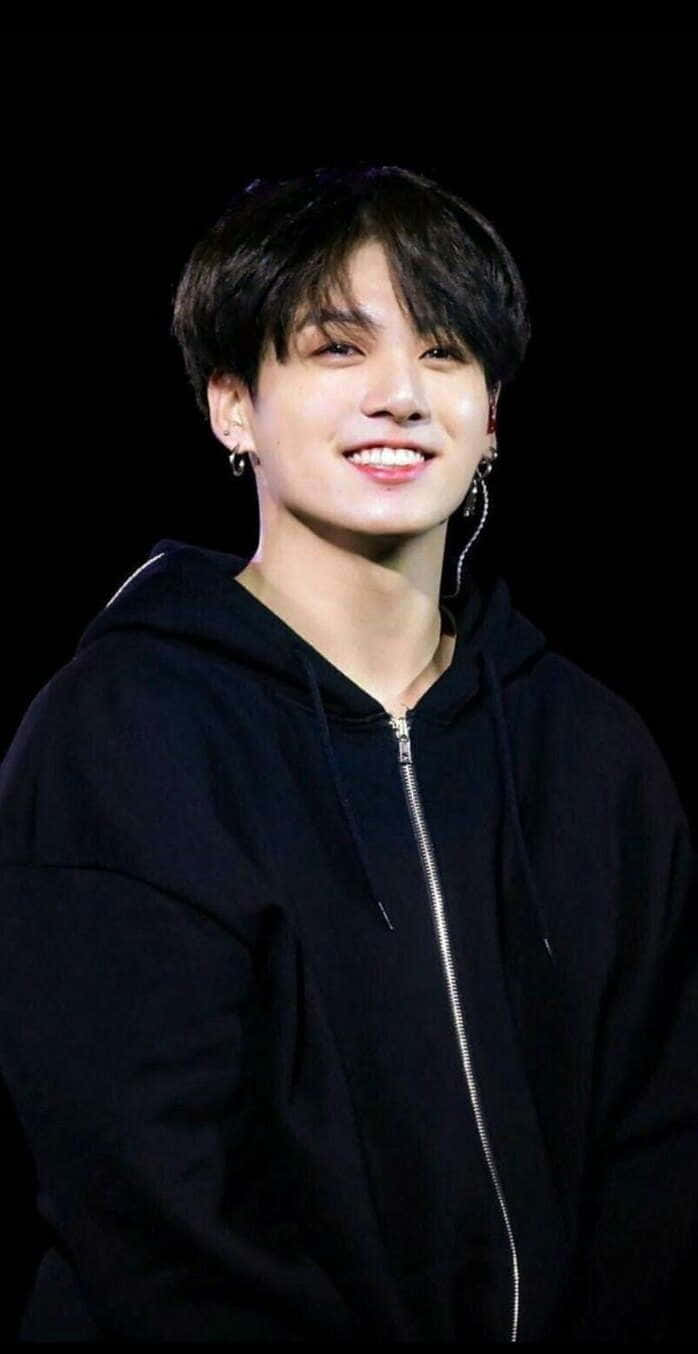 Jungkook 桌布 壁紙 — 一位深色頭髮、戴耳環的年輕男子，在黑色背景前穿著黑色拉鍊連帽衫，溫暖地微笑。