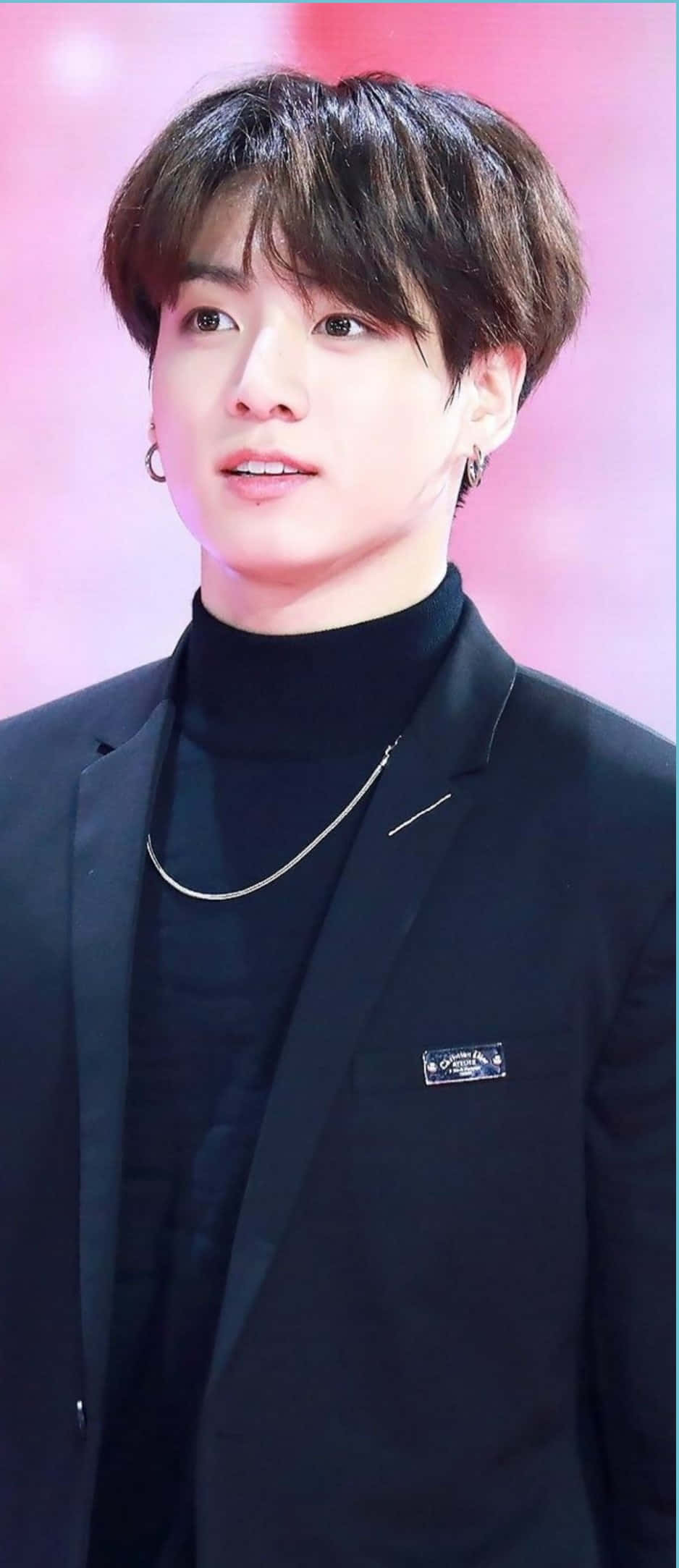 Jungkook 桌布 壁紙 — 一位深色頭髮、戴耳環的年輕男子，在柔和的粉紅色背景前穿著黑色高領毛衣和西裝外套。