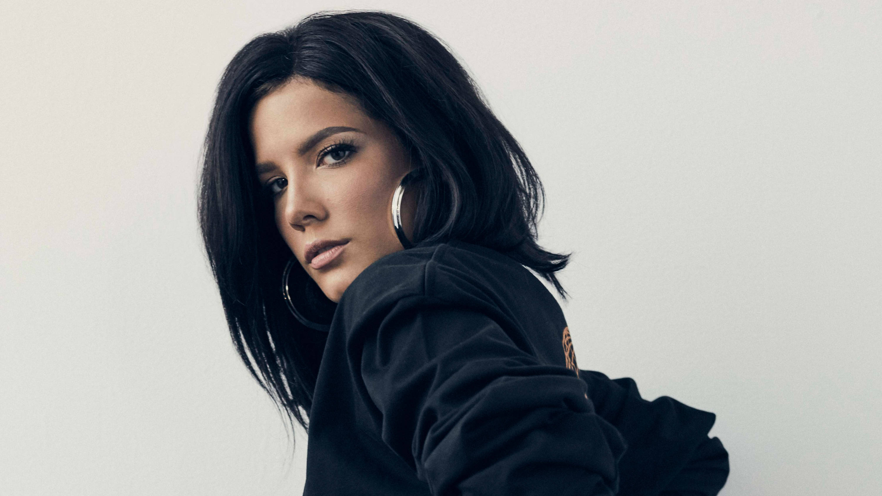 Halsey 桌布 壁紙 — 一位深色頭髮、戴著大圈耳環的女性，穿著黑色連帽衫，在純白背景上轉頭越過肩膀。