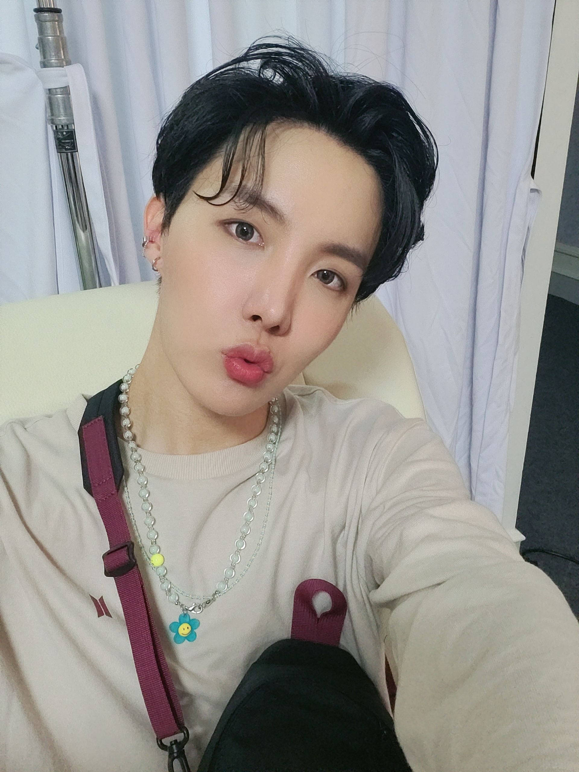 jhope 桌布 壁紙 — 一位深色頭髮、戴著珍珠項鍊的年輕人，穿著米色襯衫和紫色背包帶，做出頑皮的嘟嘴表情。