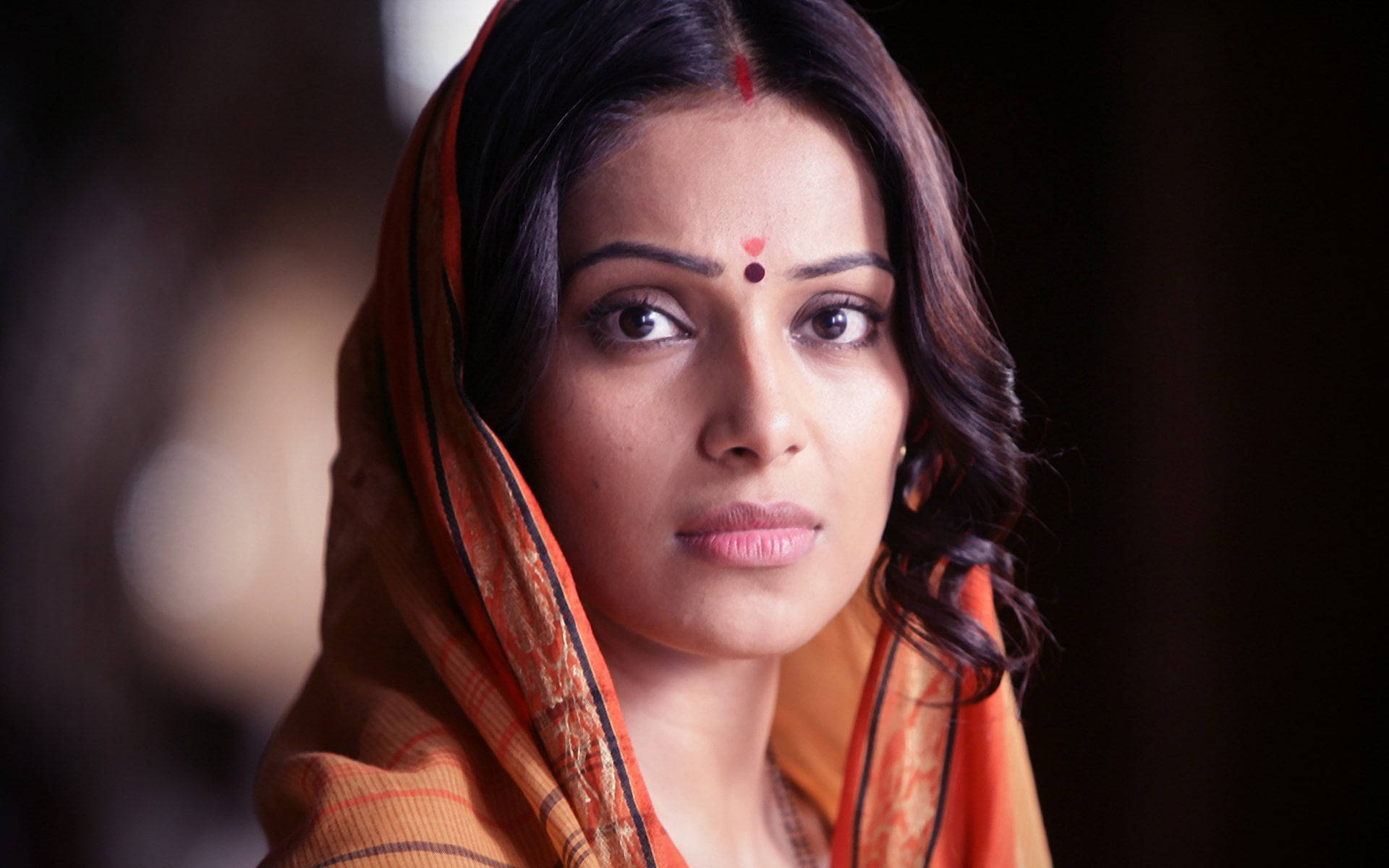 Bipasha Basu 桌布 壁紙 — 一位深色頭髮、戴著紅色眉心貼的女性，在黑暗模糊的背景前，披著橙色圖案圍巾，專注地凝視。