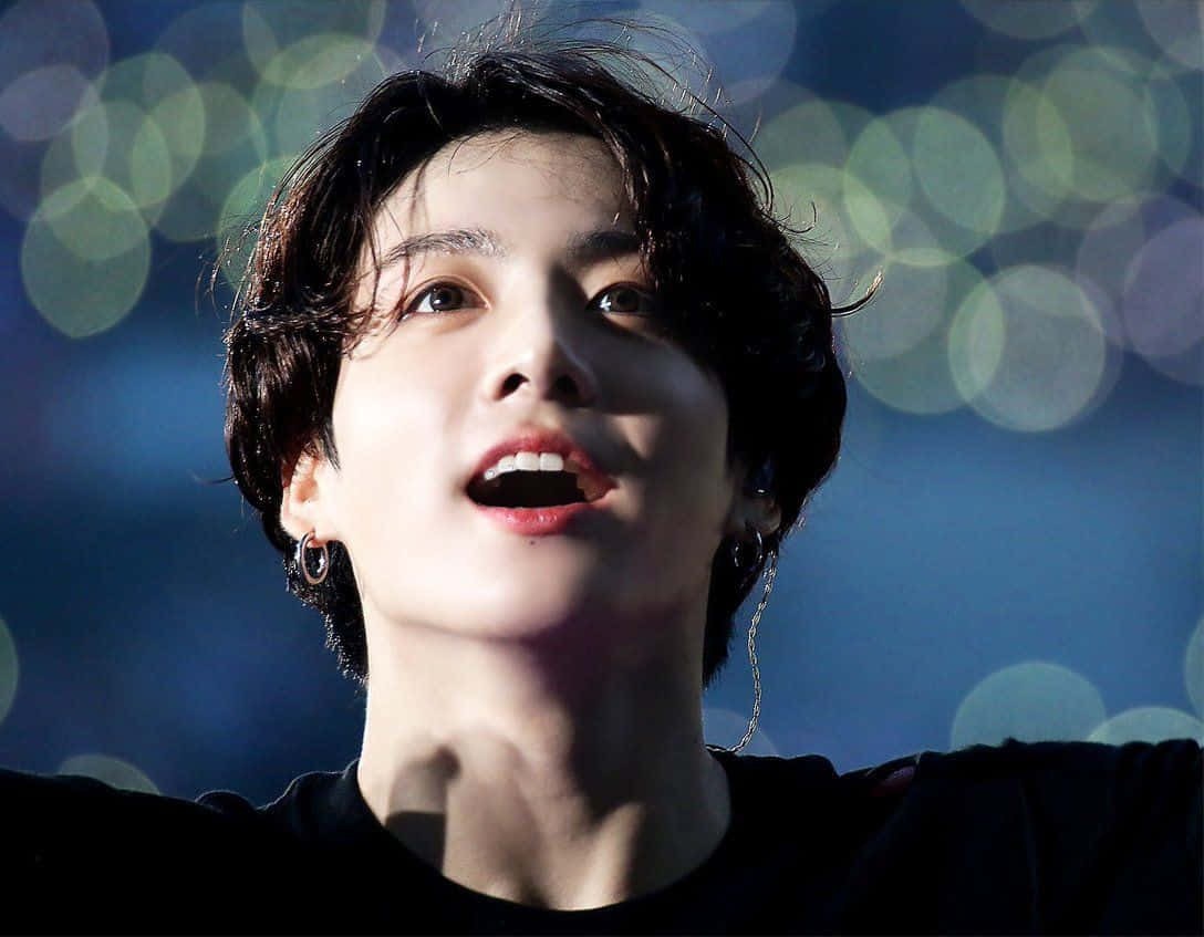 Jungkook 桌布 壁紙 — 一位深色頭髮、戴著耳環的年輕男子，在藍色散景背景下張開嘴巴向上凝視。