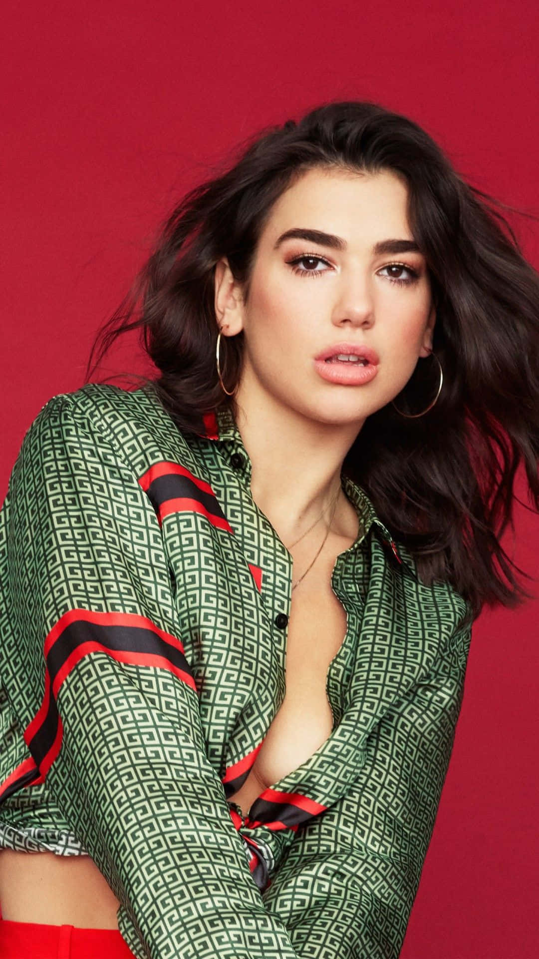 Dua Lipa 桌布 壁紙 — 一位深色頭髮、戴著金色圓環耳環的女性，身穿帶有紅色和黑色條紋的綠色圖案襯衫，背景為純紅色。