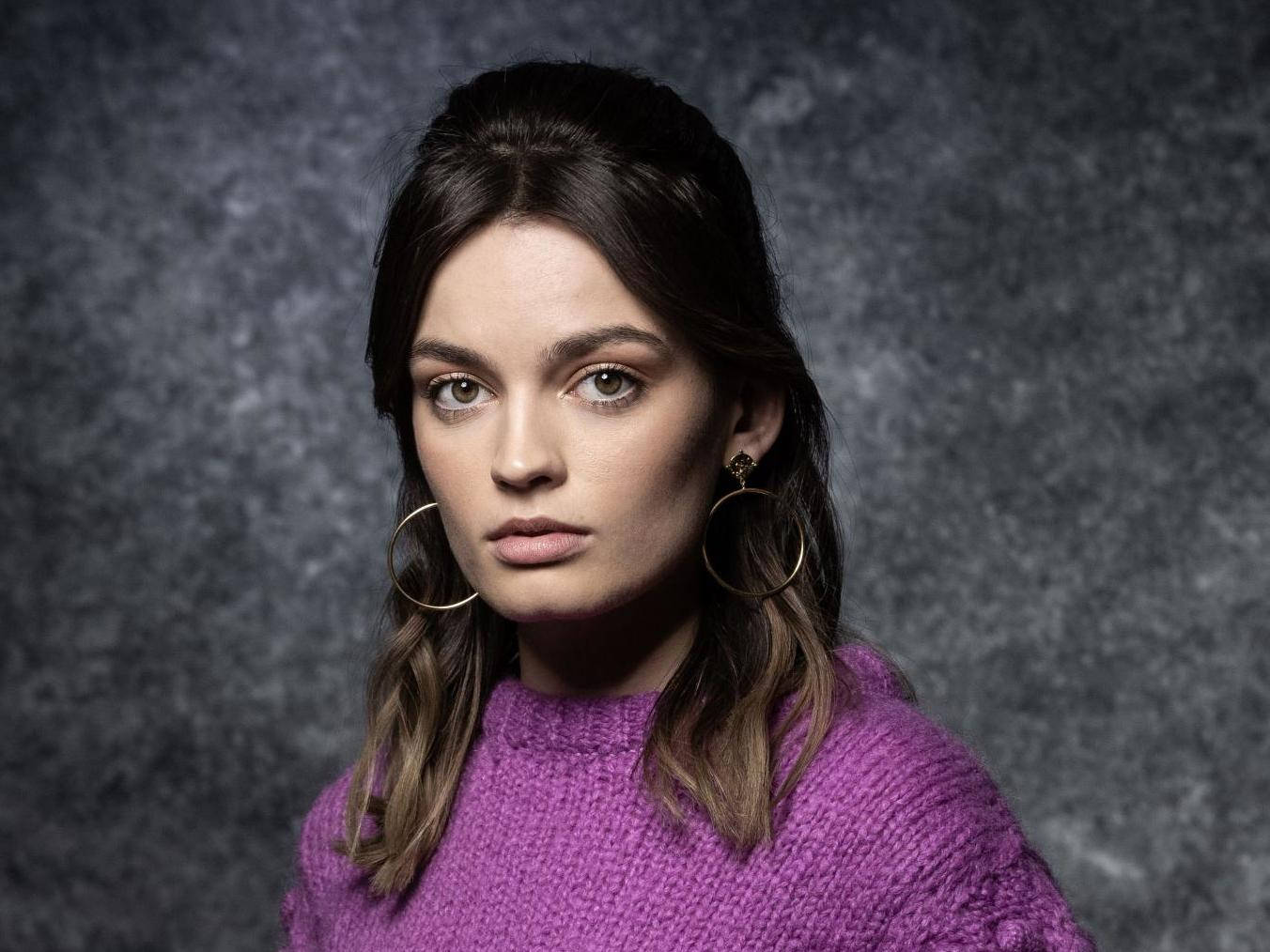maeve wiley 桌布 壁紙 — 一位深色頭髮、戴著金色圓環耳環的女性，在灰色工作室背景下穿著一件有紋理的紫色毛衣。