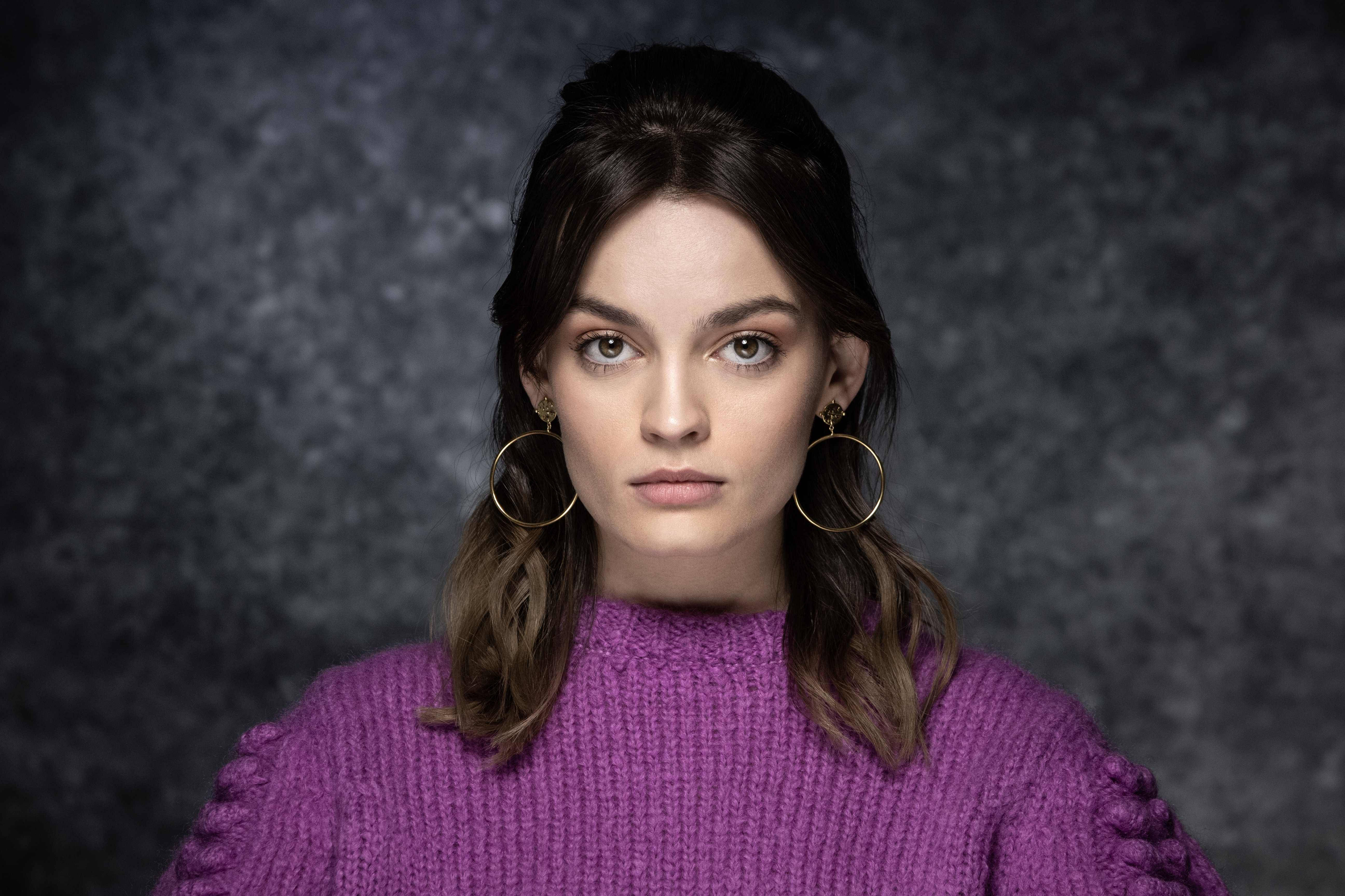 Emma 桌布 壁紙 — 一位深色頭髮、戴著金色圓環耳環的女性，在深灰色背景下穿著一件有紋理的紫色毛衣。