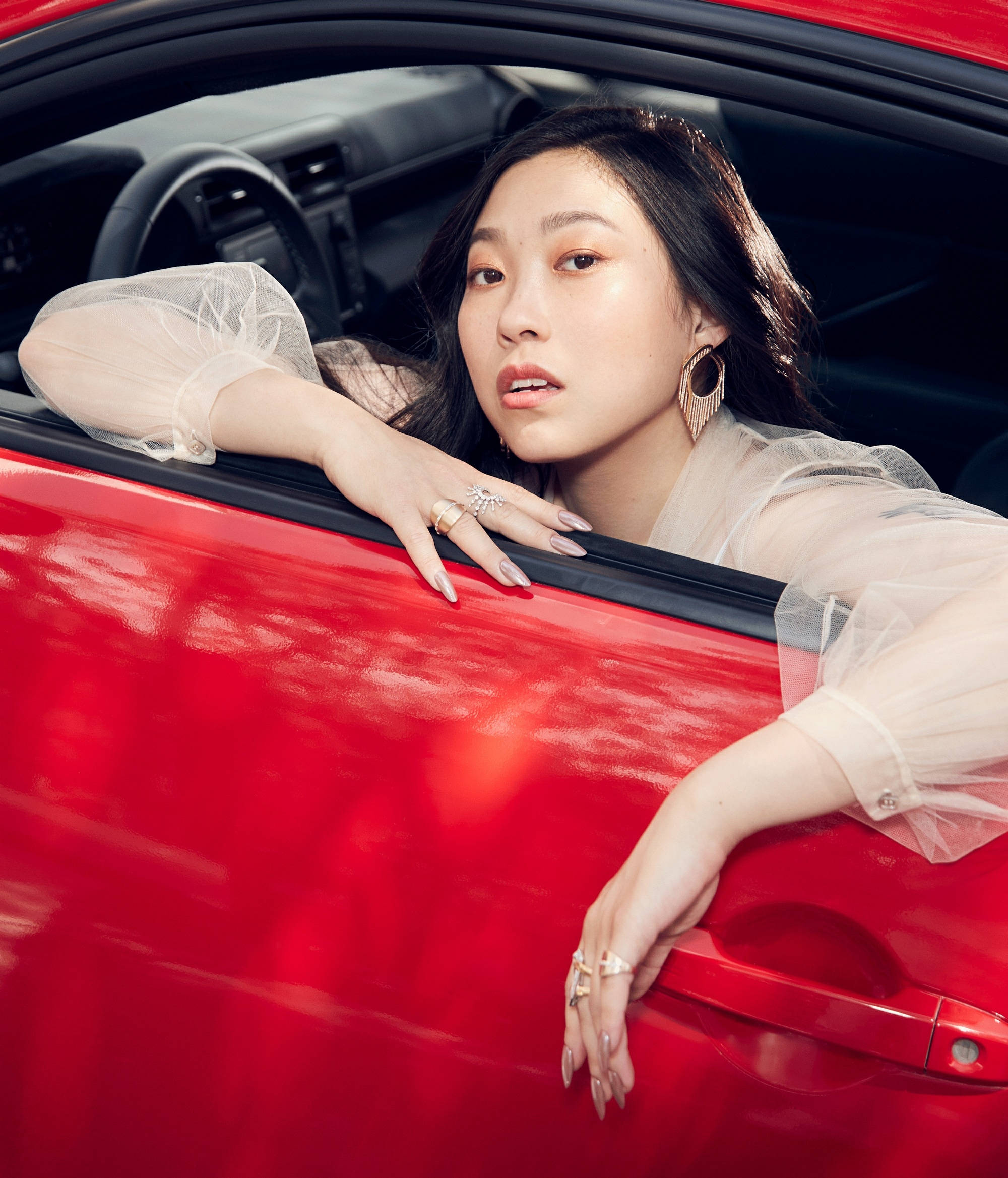 Awkwafina 桌布 壁紙 — 一位深色頭髮、戴著金色耳環的女性，從一輛亮紅色車窗探出身子，身穿透視襯衫和戒指。