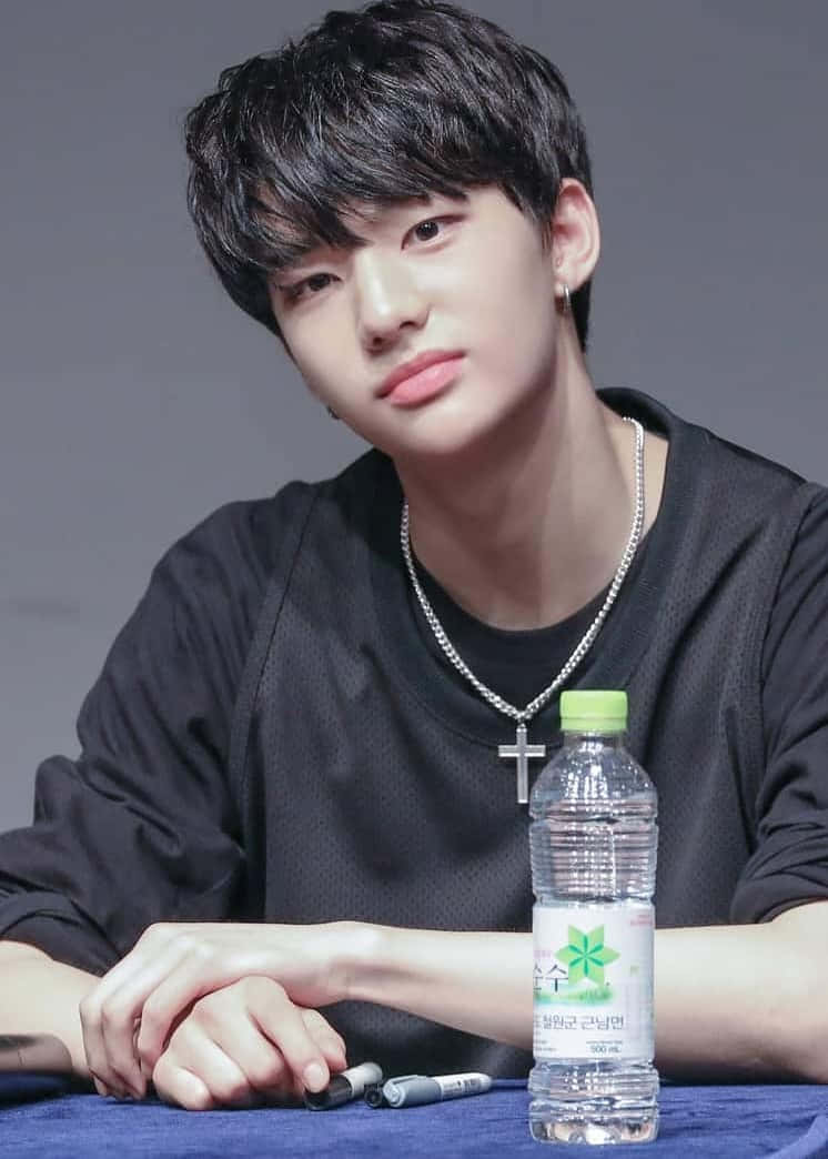 hyunjin 桌布 壁紙 — 一位深色頭髮、戴著銀色十字架項鍊的年輕男子坐在一張放有水壺和筆的桌子旁。