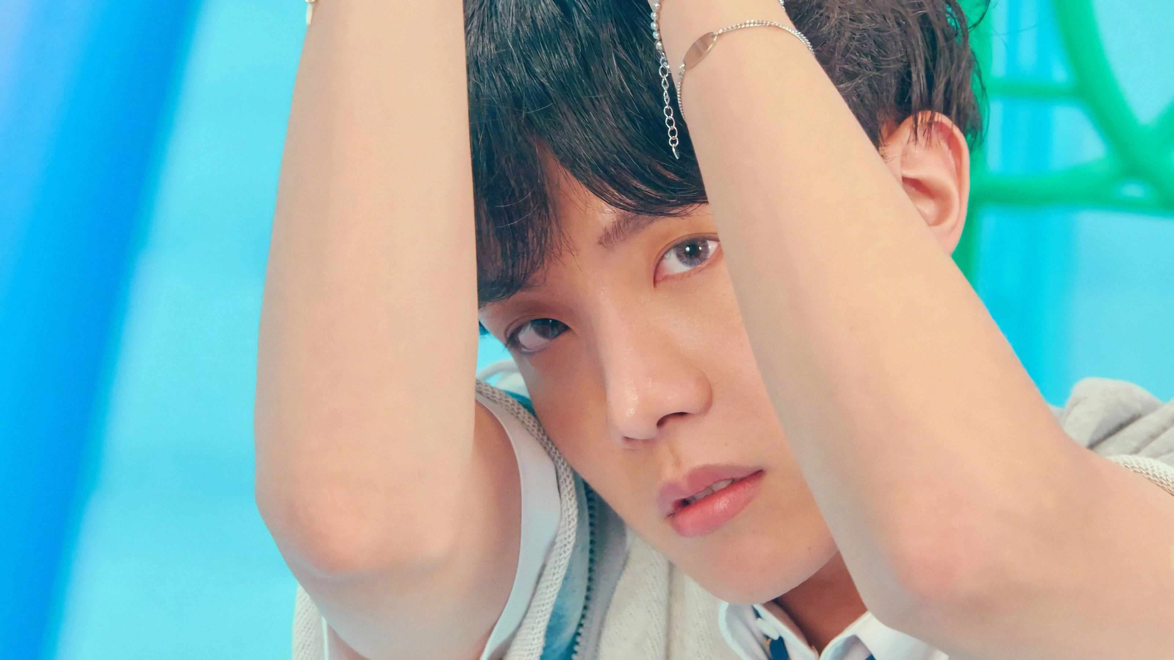 j-hope 桌布 壁紙 — 一位深色頭髮、戴著銀鏈手鐲的年輕男子，在充滿活力的藍色和綠色背景前，雙臂高舉過頭，強烈地凝視著鏡頭。