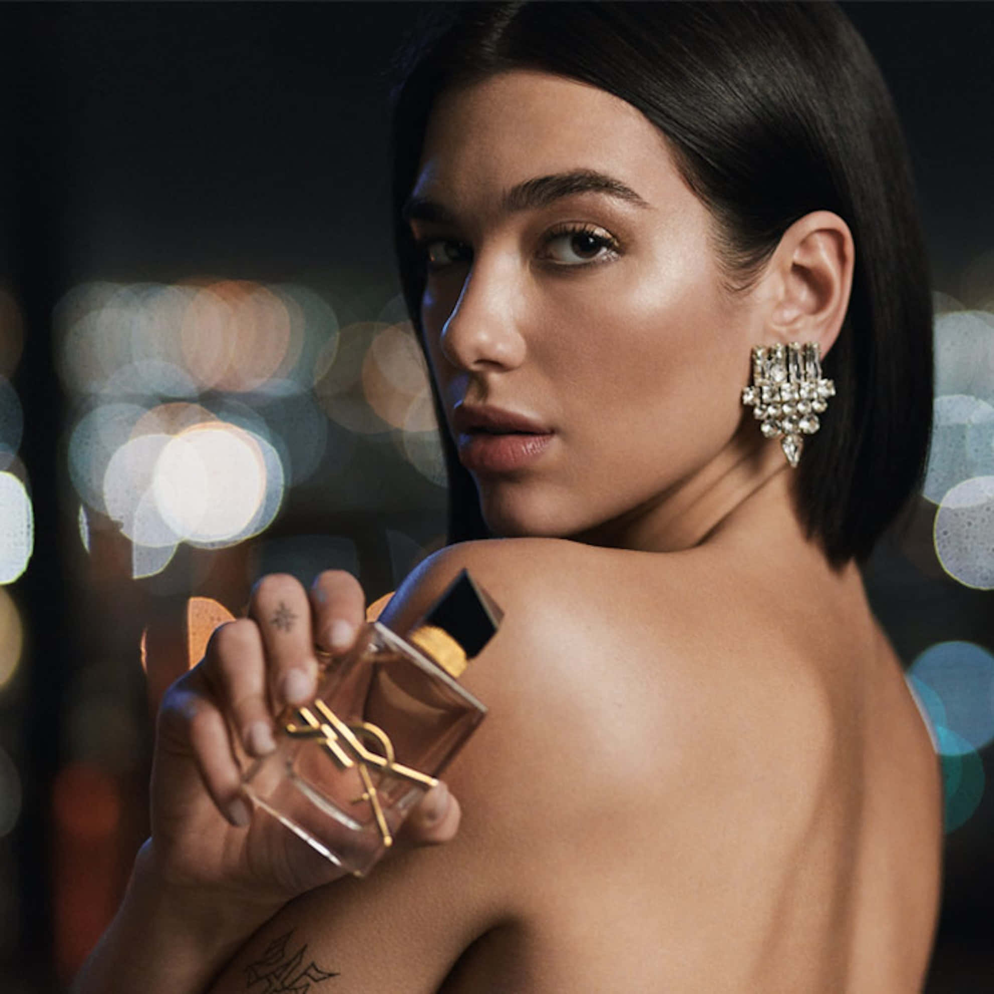 YSL 桌布 壁紙 — 一位深色頭髮、戴著鑽石耳環的女性，在模糊的城市夜景背景下，將香水瓶貼在裸露的肩膀上。