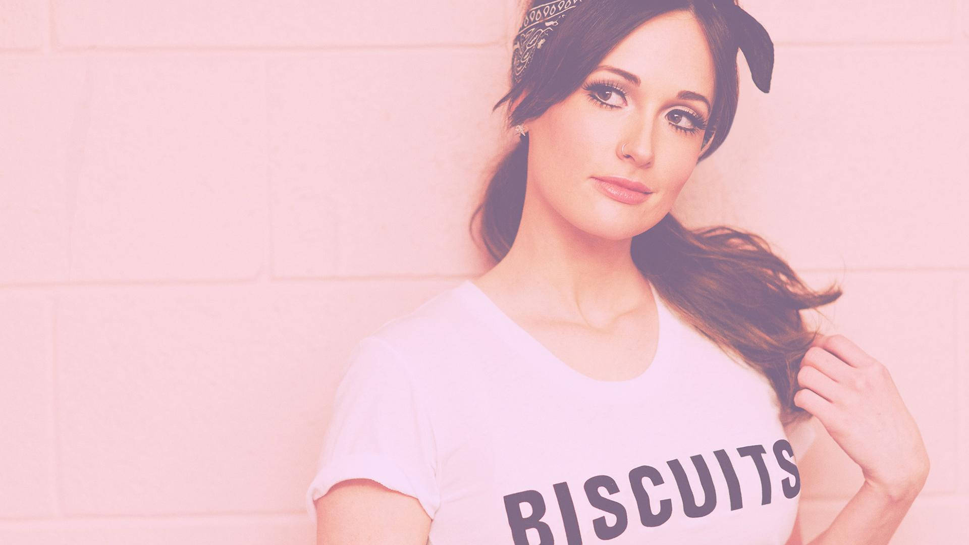 Kacey Musgraves 桌布 壁紙 — 一位深色頭髮、戴頭巾的女性，身穿印有biscuits字樣的白色T恤，靠著粉紅磚牆。