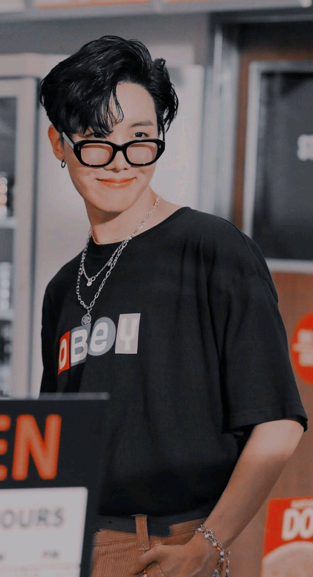 jhope 桌布 壁紙 — 一位深色頭髮、戴黑色眼鏡的年輕男子，穿著黑色 'OBEY' T恤和層疊項鍊微笑。