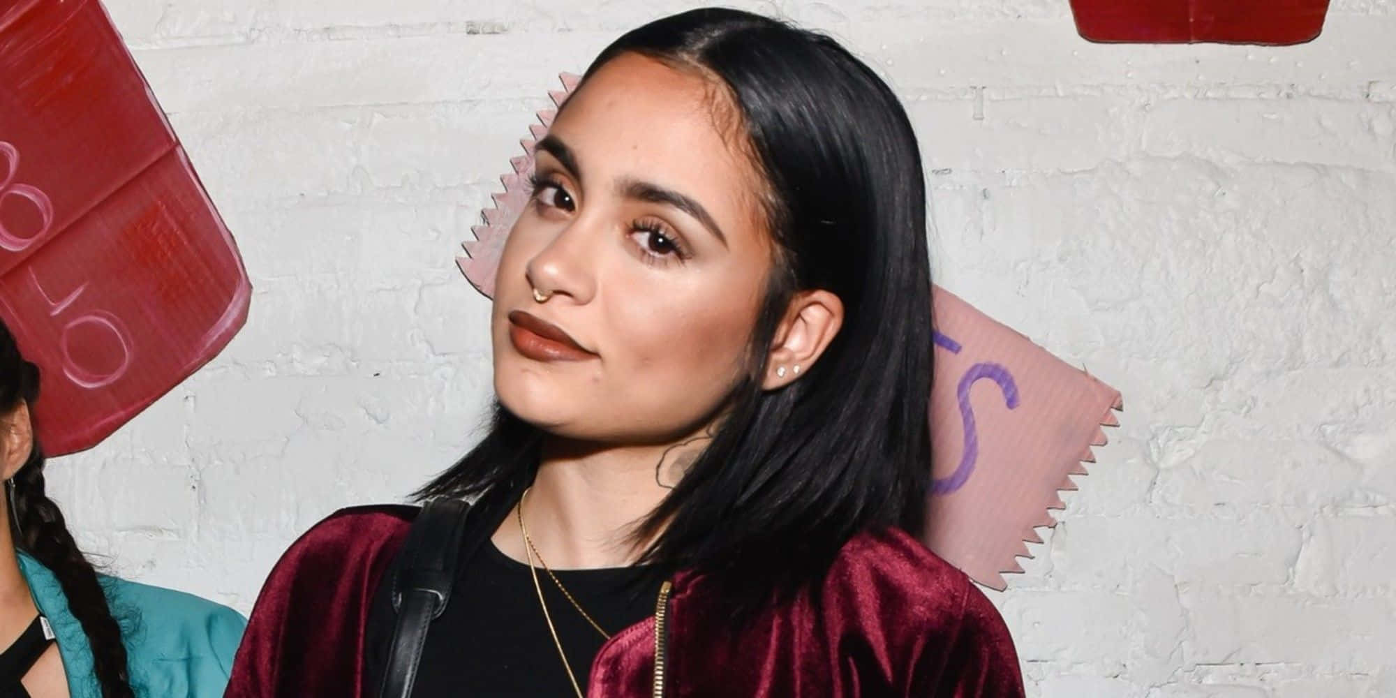 kehlani 桌布 壁紙 — 一位深色頭髮、戴鼻環的年輕女性，身穿栗色天鵝絨外套，背景是帶有彩色塗鴉的白色磚牆。