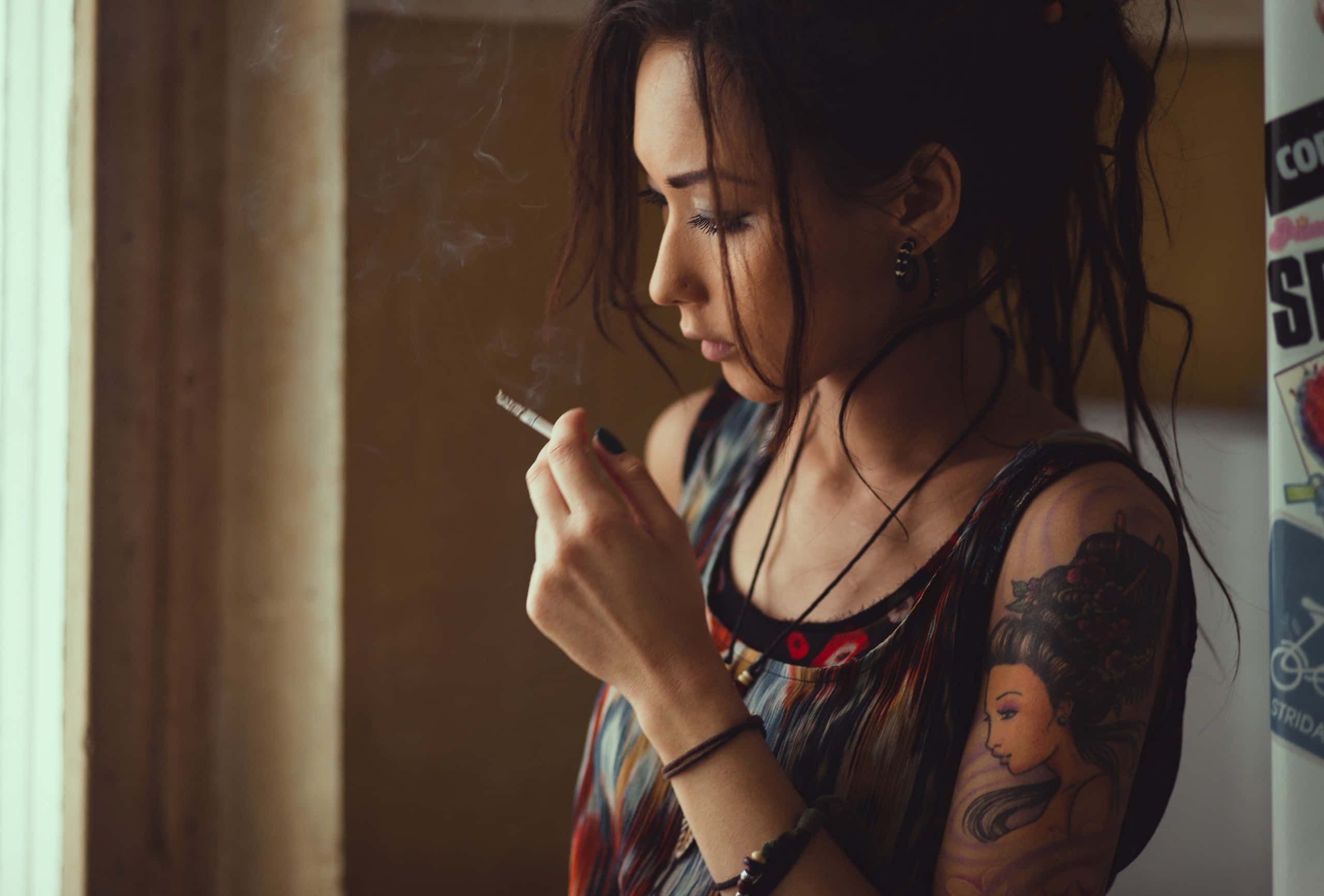 Girl Smoking 桌布 壁紙 — 一位深色頭髮、手臂有刺青的女人，在昏暗的房間裡靠近窗戶抽菸。