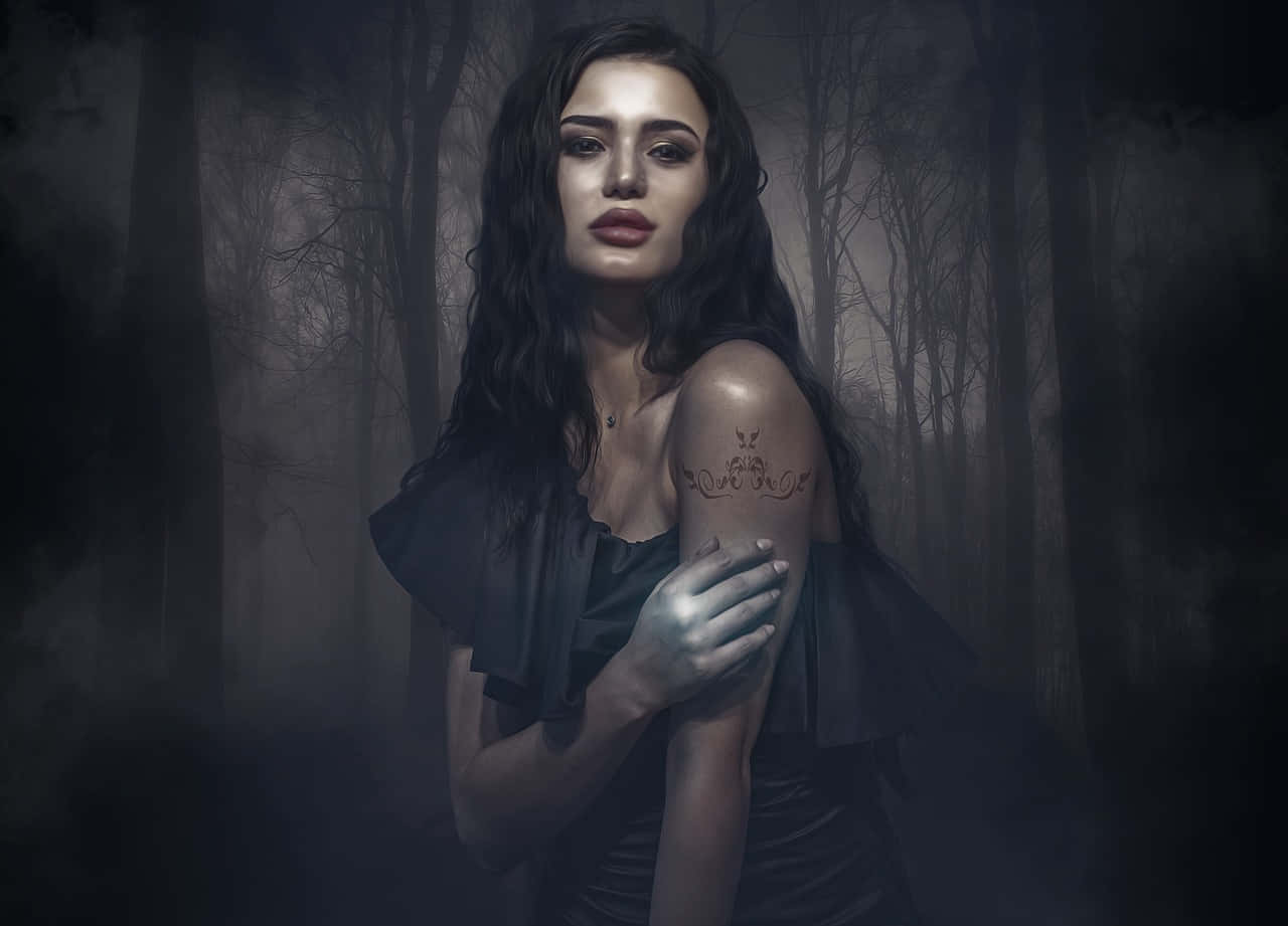 Gothic Fashion 桌布 壁紙 — 一位深色頭髮、手臂有刺青的女性，站在霧氣瀰漫、陰暗的森林中，周圍是光禿禿的樹木。