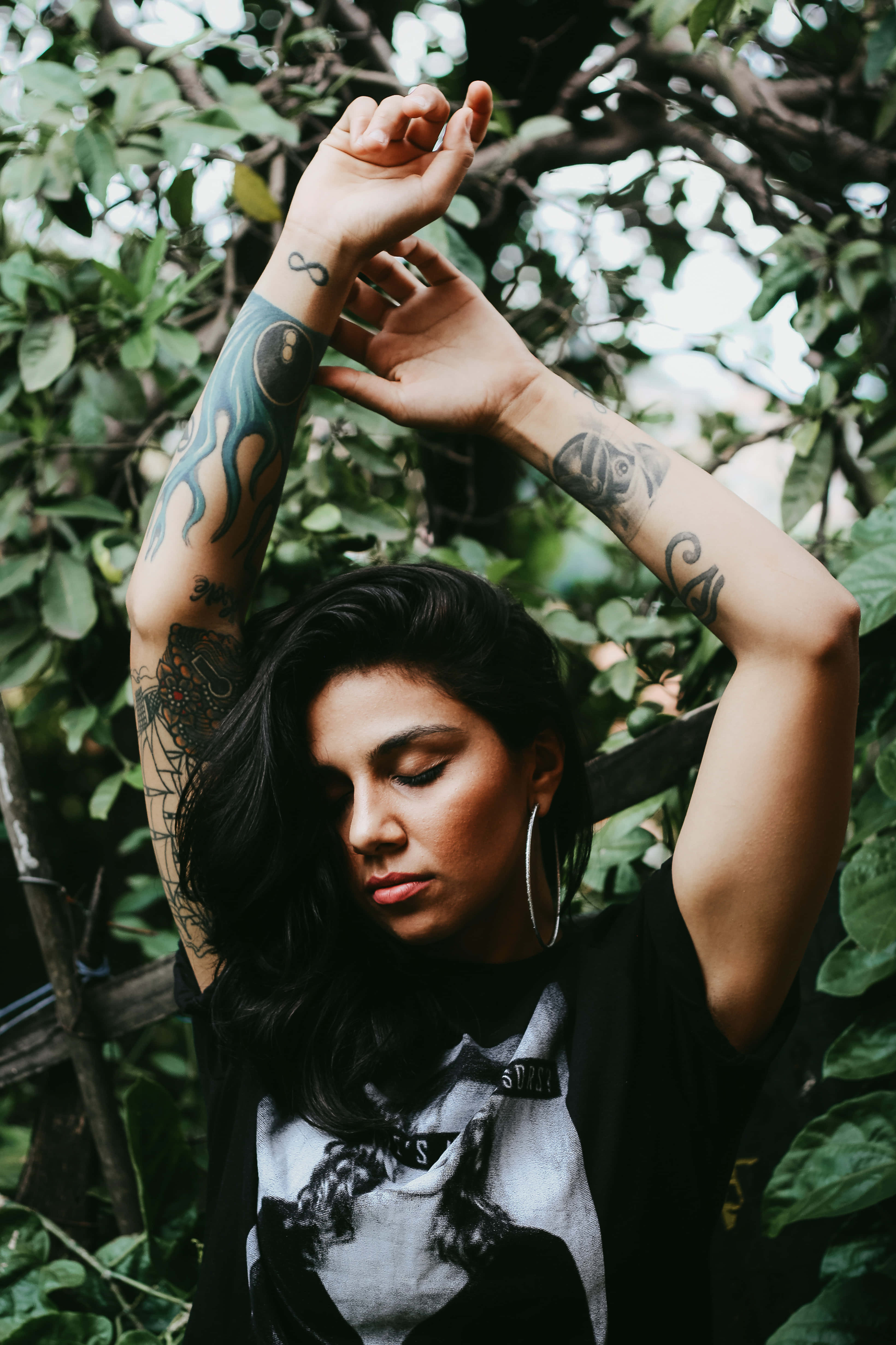 Tattoo Girl 桌布 壁紙 — 一位深色頭髮、手臂有刺青的女性，在寧靜的戶外環境中，於茂密的綠葉間舉起雙手。