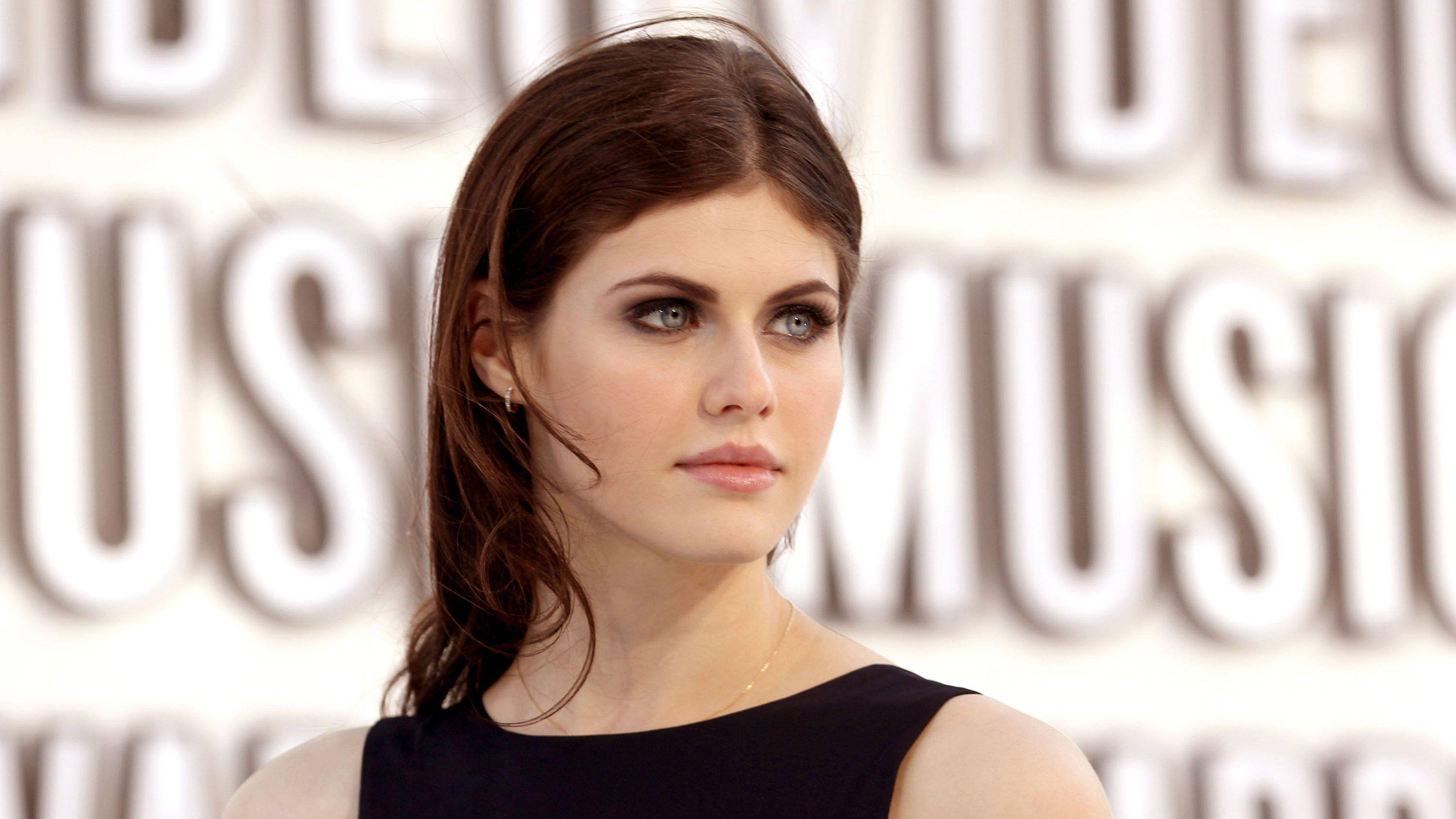 Alexandra Daddario 4K 桌布 壁紙 — 一位深色頭髮、擁有引人注目藍眼睛的女性，在一個帶有大字母的模糊白色背景前若有所思地凝視。