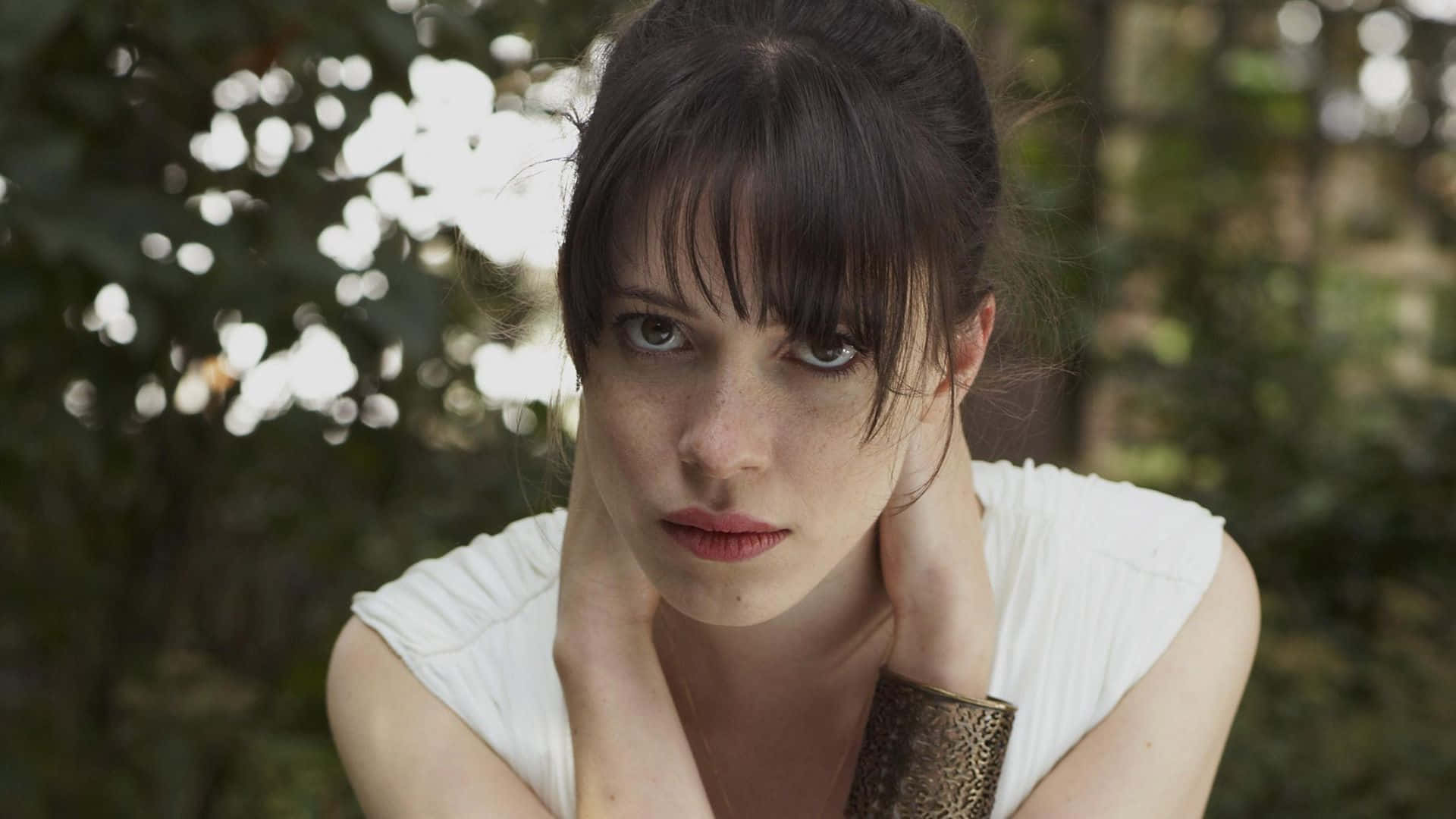 Rebecca Hall 桌布 壁紙 — 一位深色頭髮、有雀斑的女性在陽光明媚的森林中，雙手放在脖子上，專注地凝視著相機。